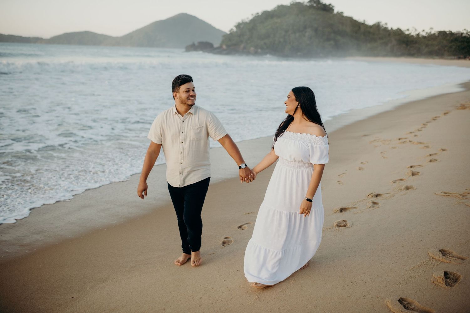 Couple walking hand in hand along a scenic beach with mountains in background.