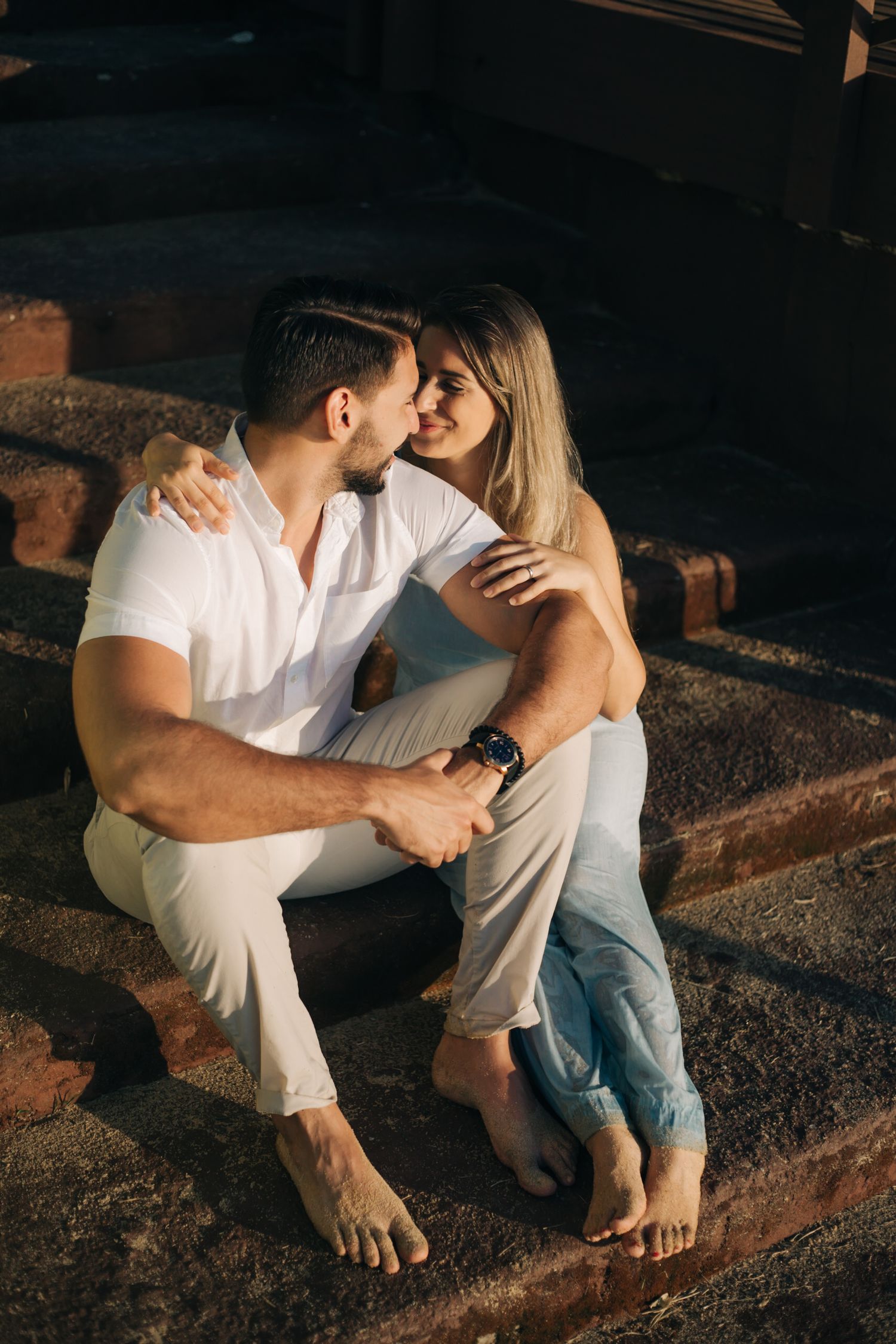 A romantic couple sits barefoot on stone steps at sunset wearing white and denim casual outfits.