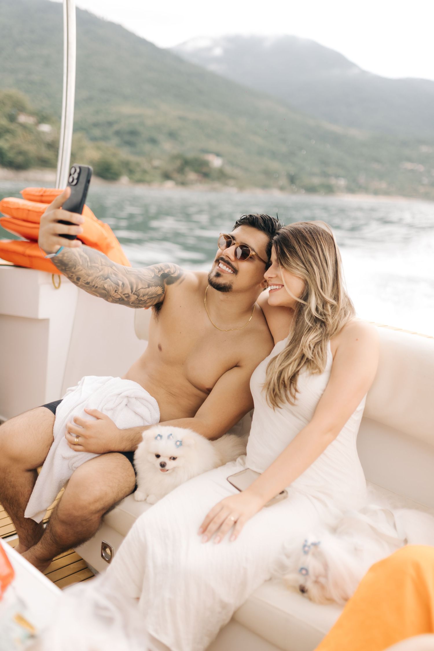 Couple taking selfie on boat with small white dog and mountain scenery in background.