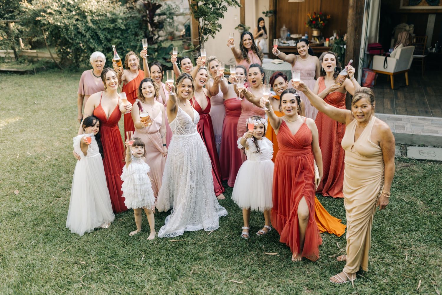 Large bridal party poses together outdoors wearing coordinated pink and coral formal attire.