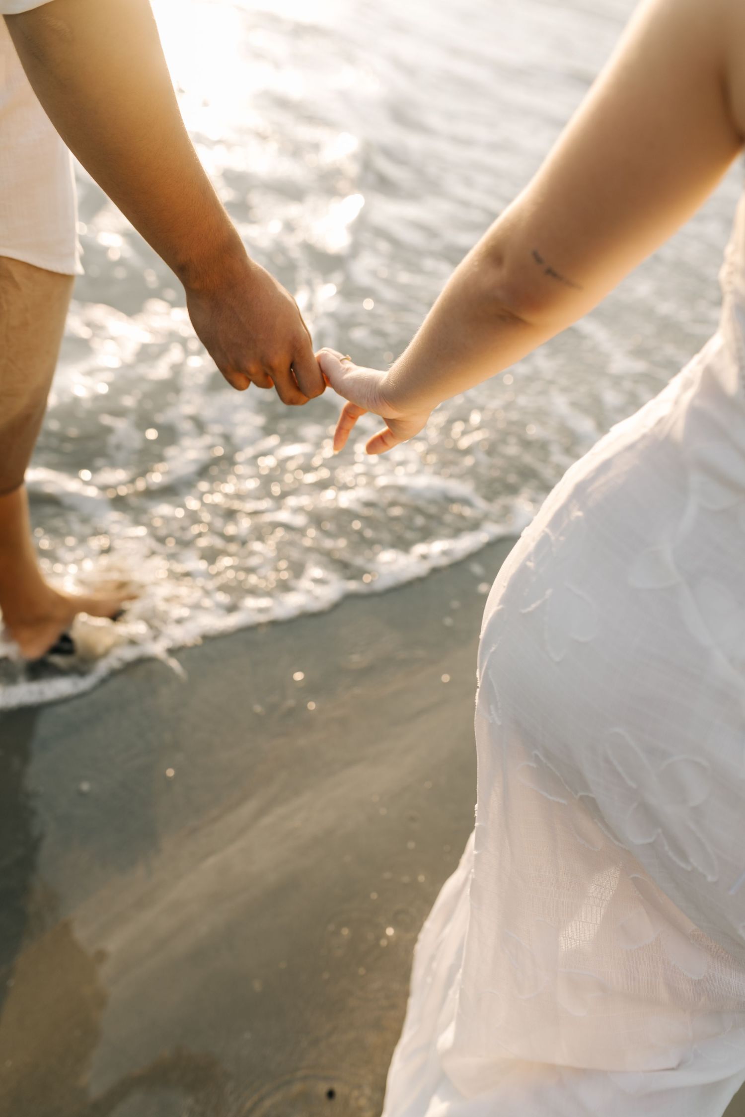 Couple holding hands while standing in shallow ocean waves during sunset.