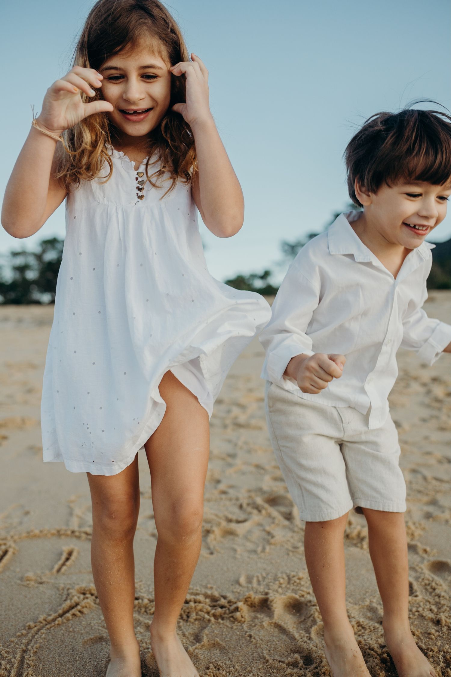 Playful kids in white clothing crouch and laugh on a sandy beach.