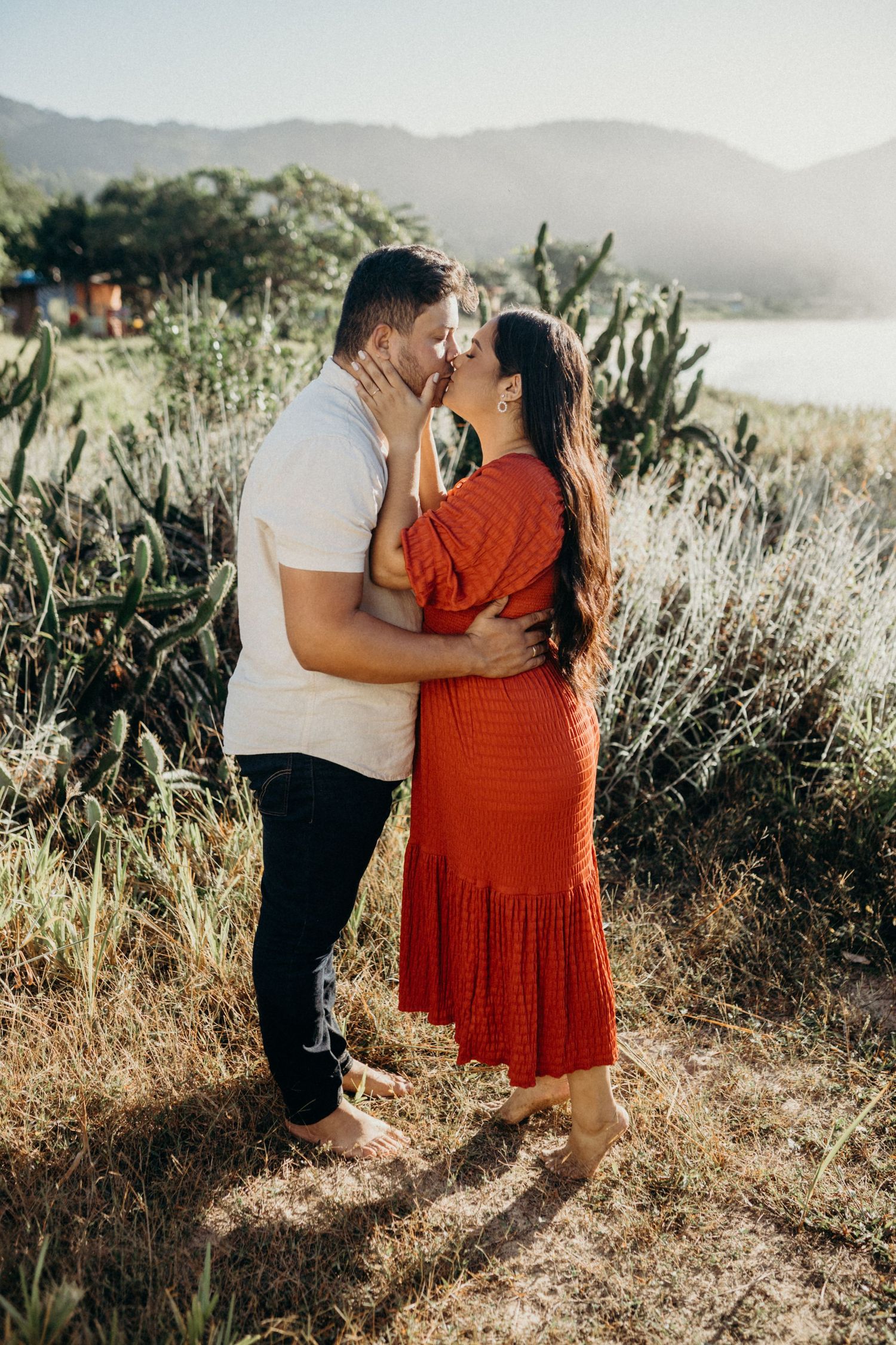 Romantic couple embracing in golden sunlit field with mountains in background during outdoor photo session.