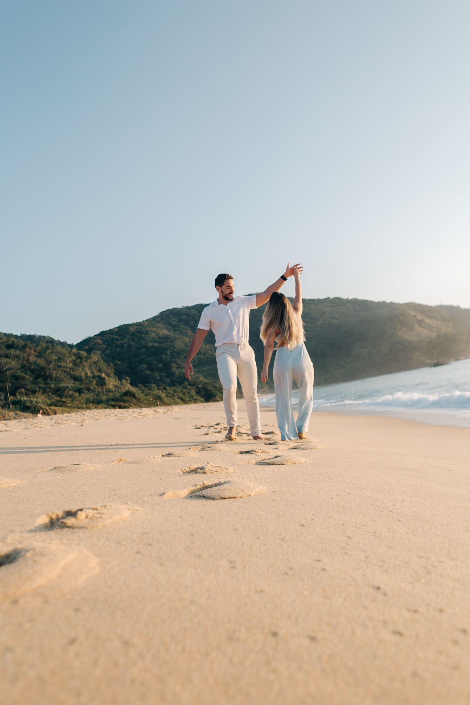 Couple holding hands while walking along an empty beach with mountain backdrop.
