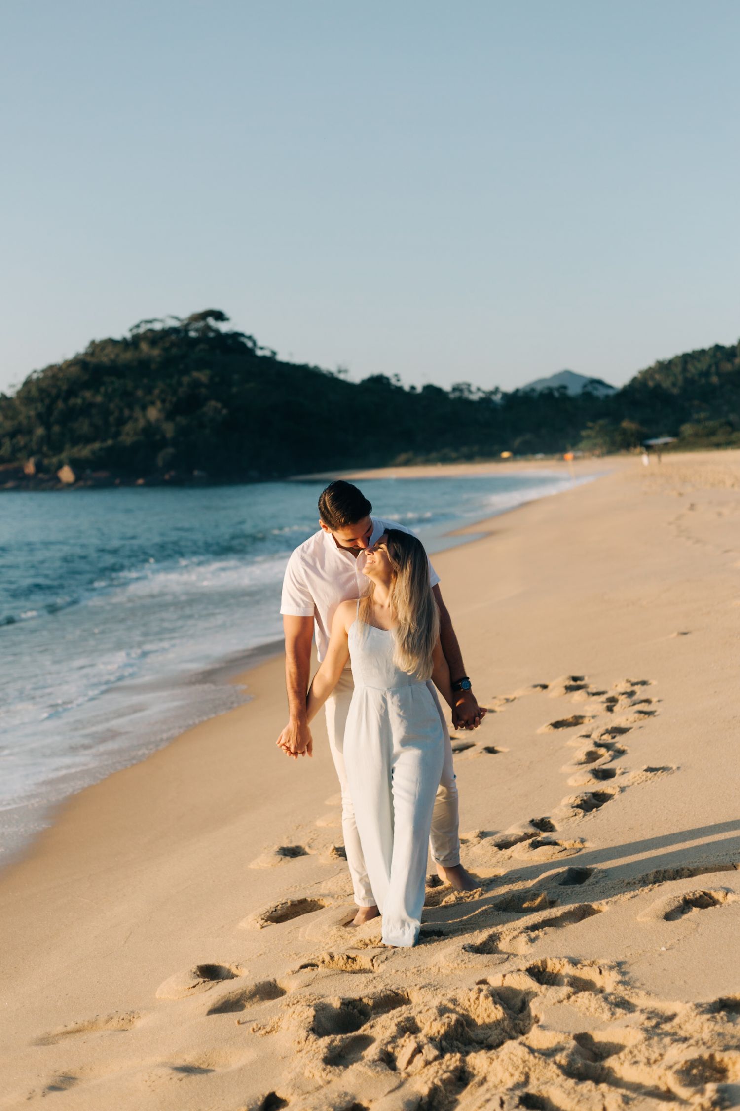 Intimate moment between couple in white attire walking on beach with ocean waves nearby.