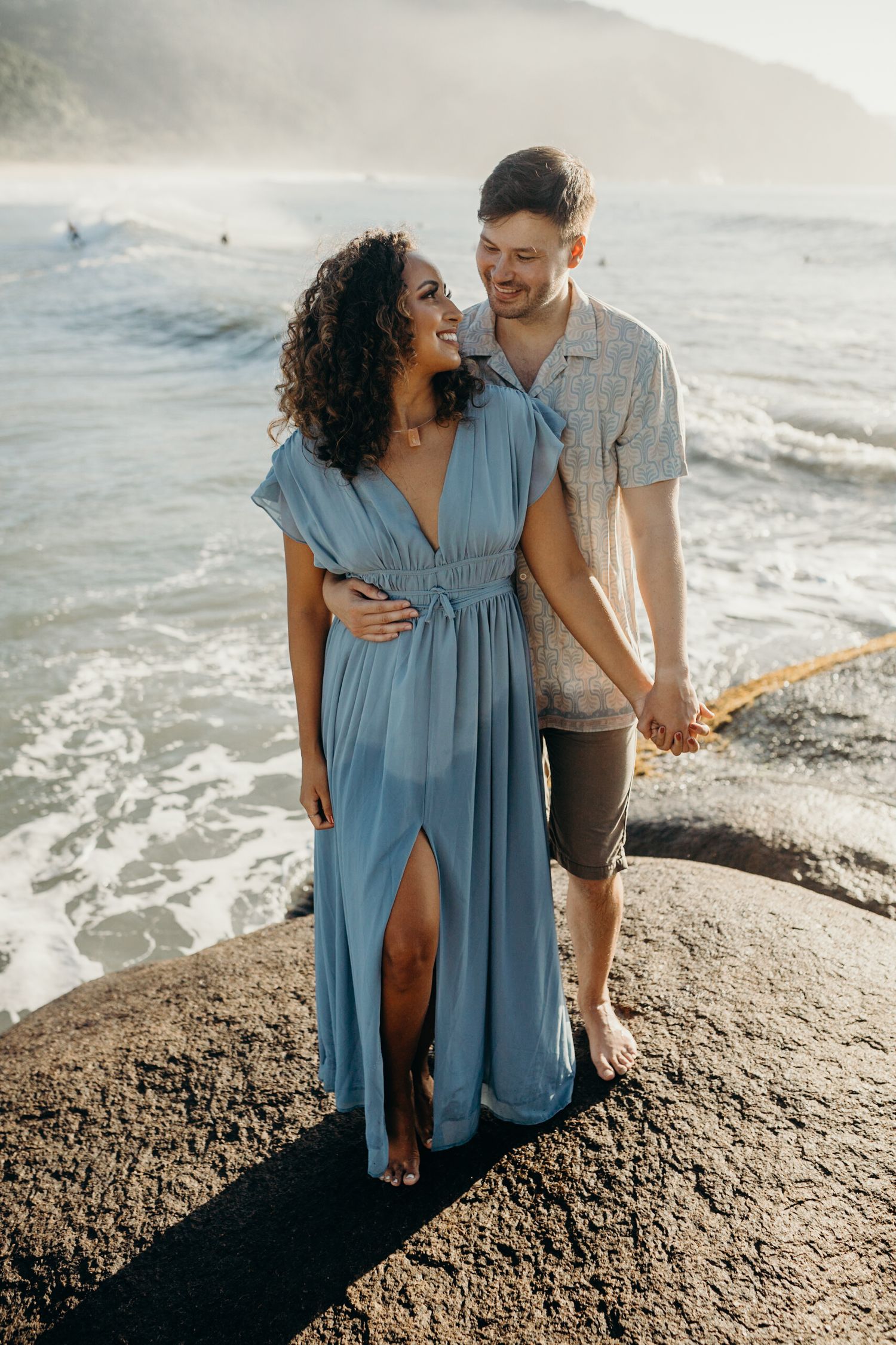 Couple standing on rocky outcrop overlooking misty ocean coastline at sunset.