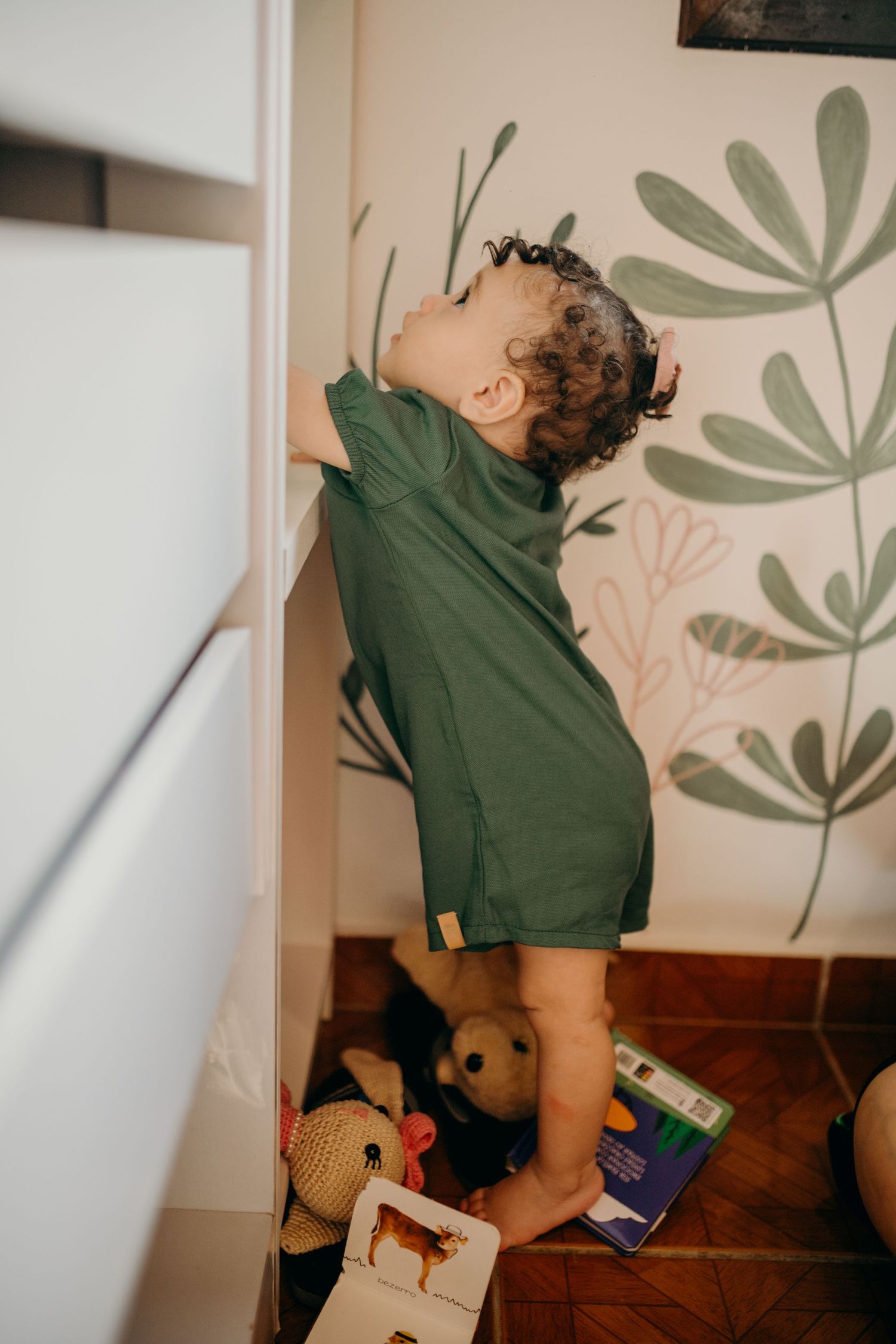 Small child in green outfit reaching up towards wall with floral decals in nursery.