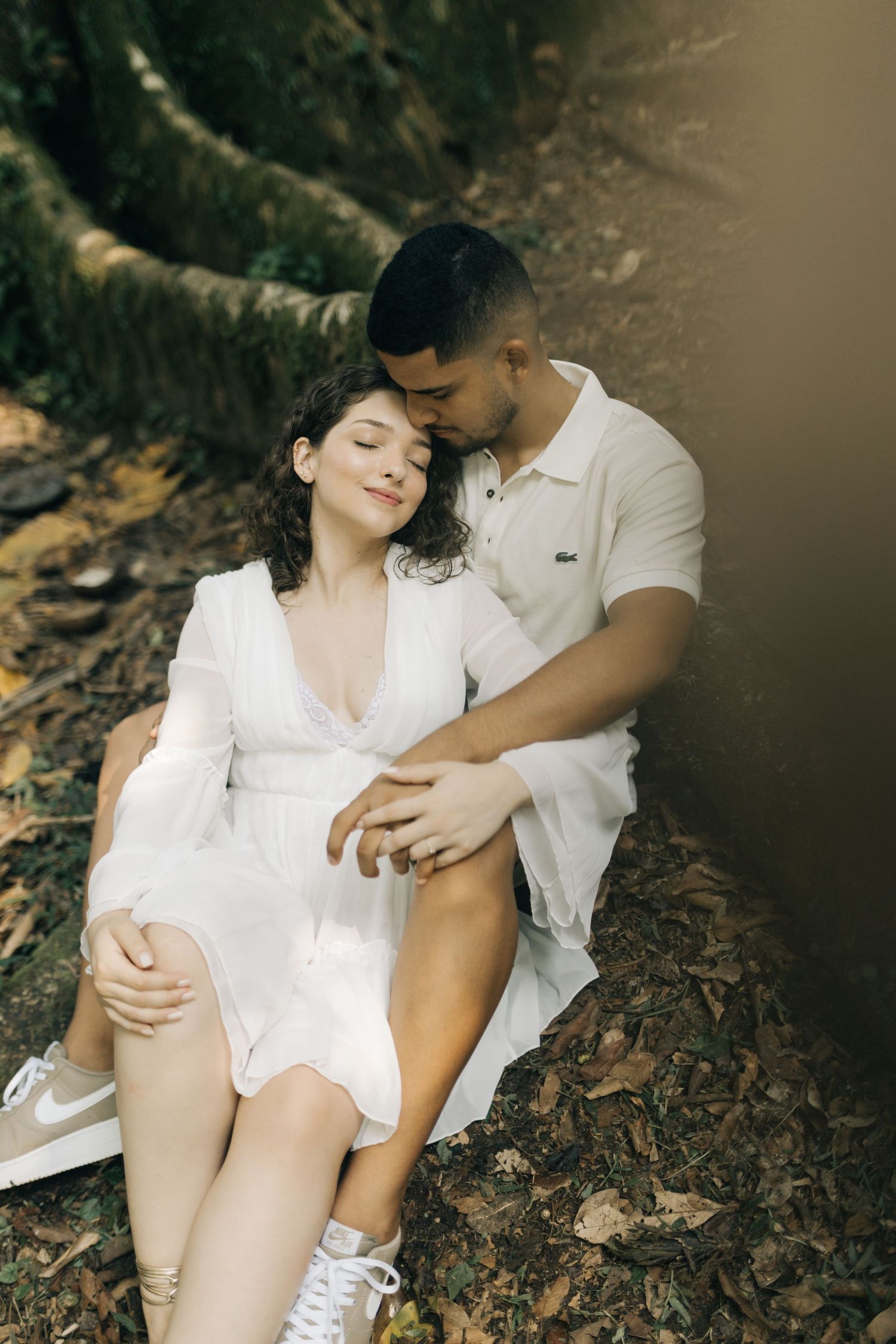 A romantic couple rests intimately against a tree in a forest setting.