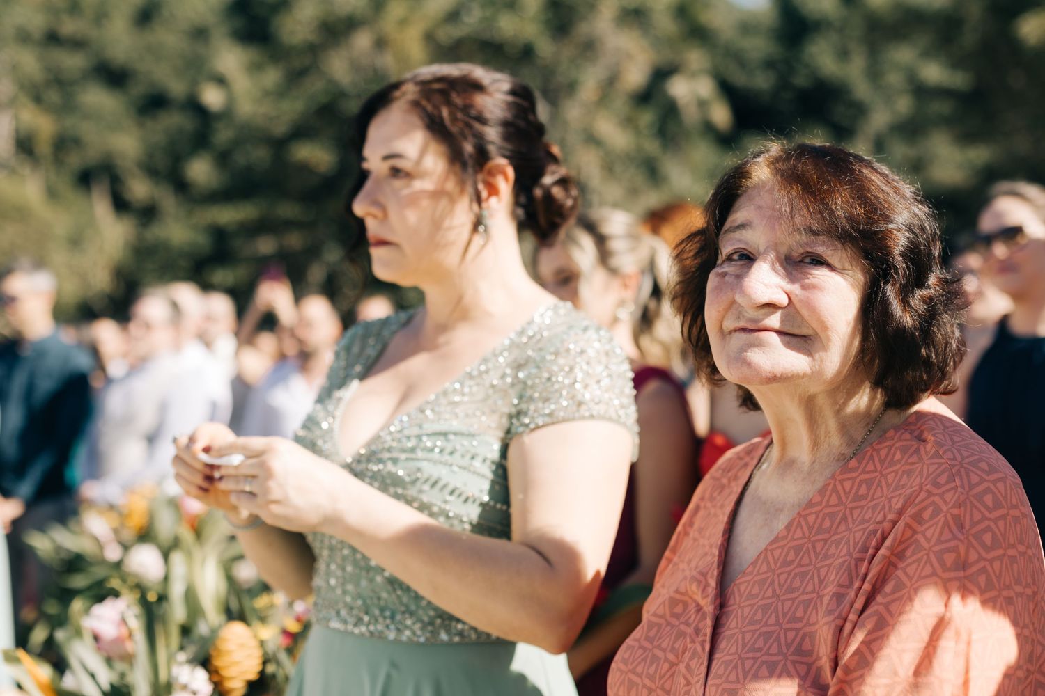 Wedding guests watching ceremony while standing outdoors in formal attire.