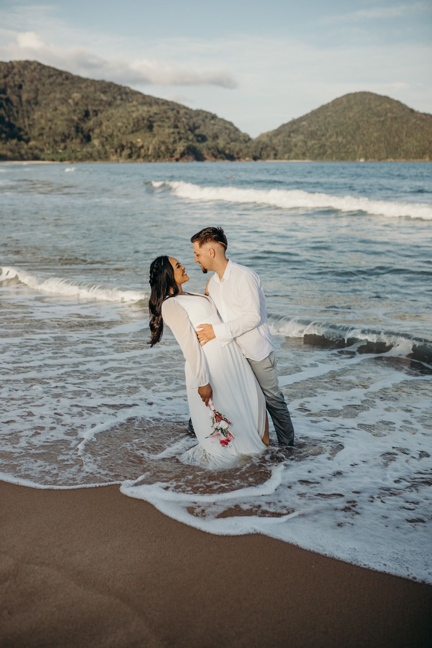 Playful couple gets splashed by ocean waves while wearing white outfits during beach photoshoot.