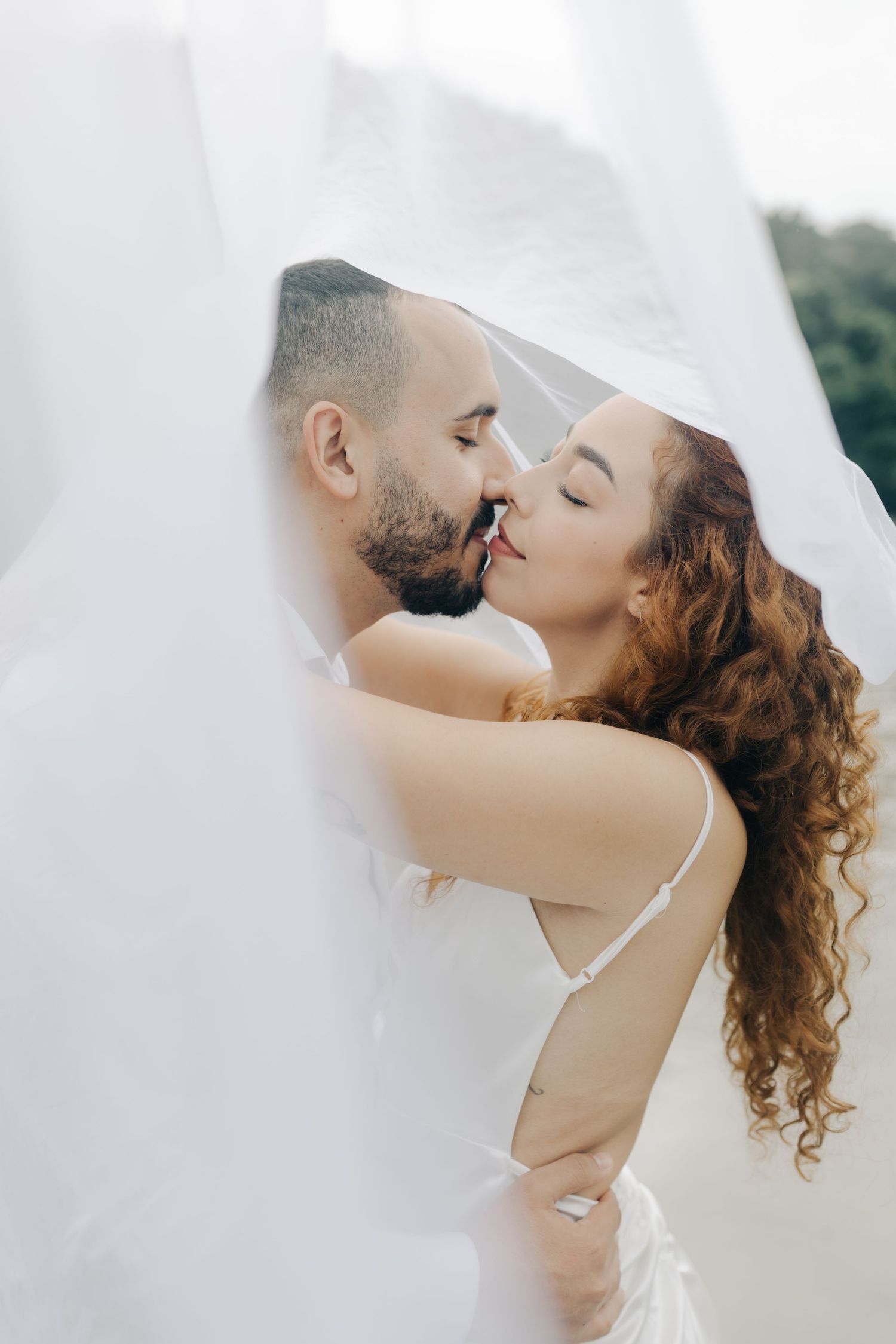 Romantic wedding couple sharing intimate moment beneath flowing veil on beach.