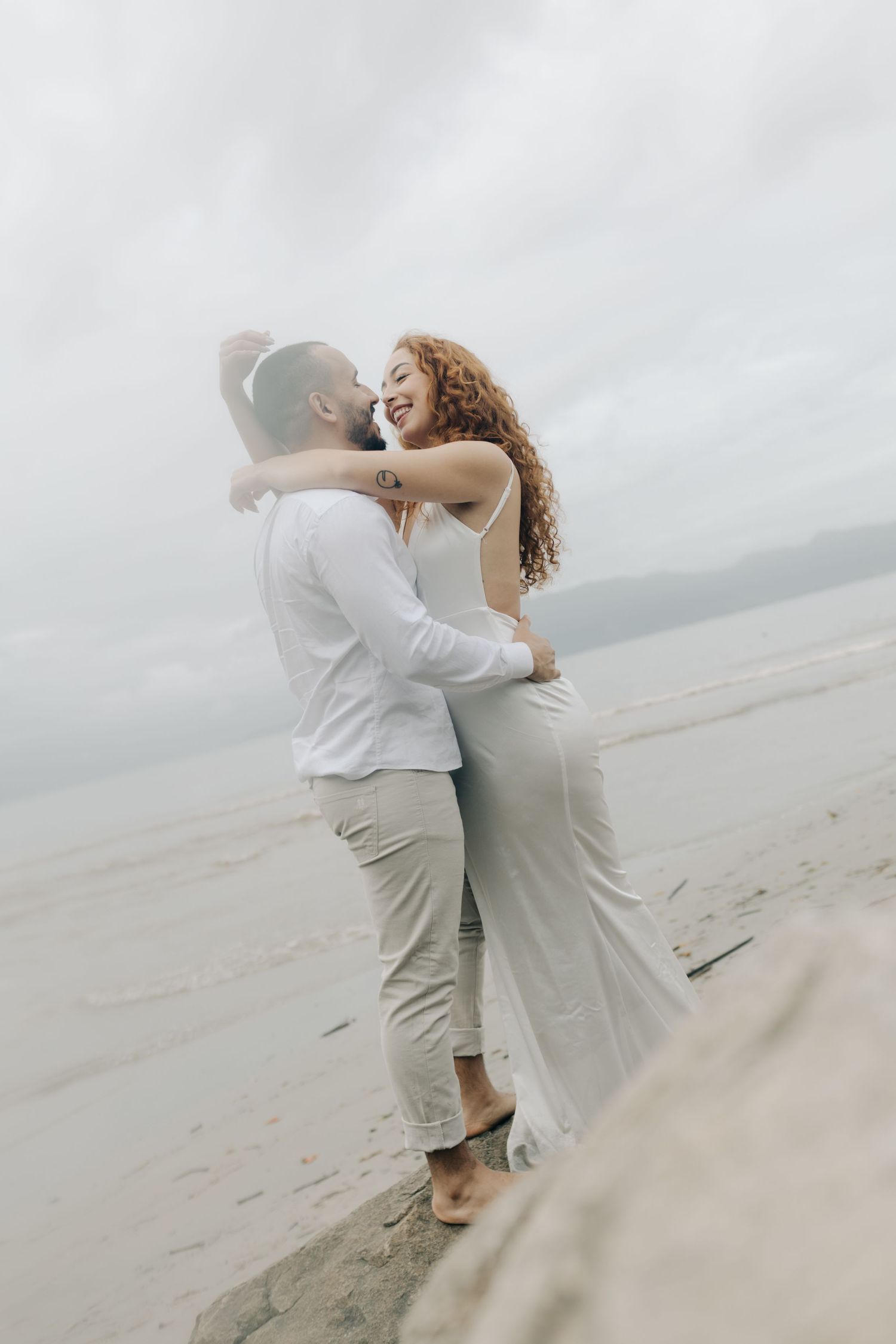 A couple in white clothing embraces tenderly on a misty beach shoreline.