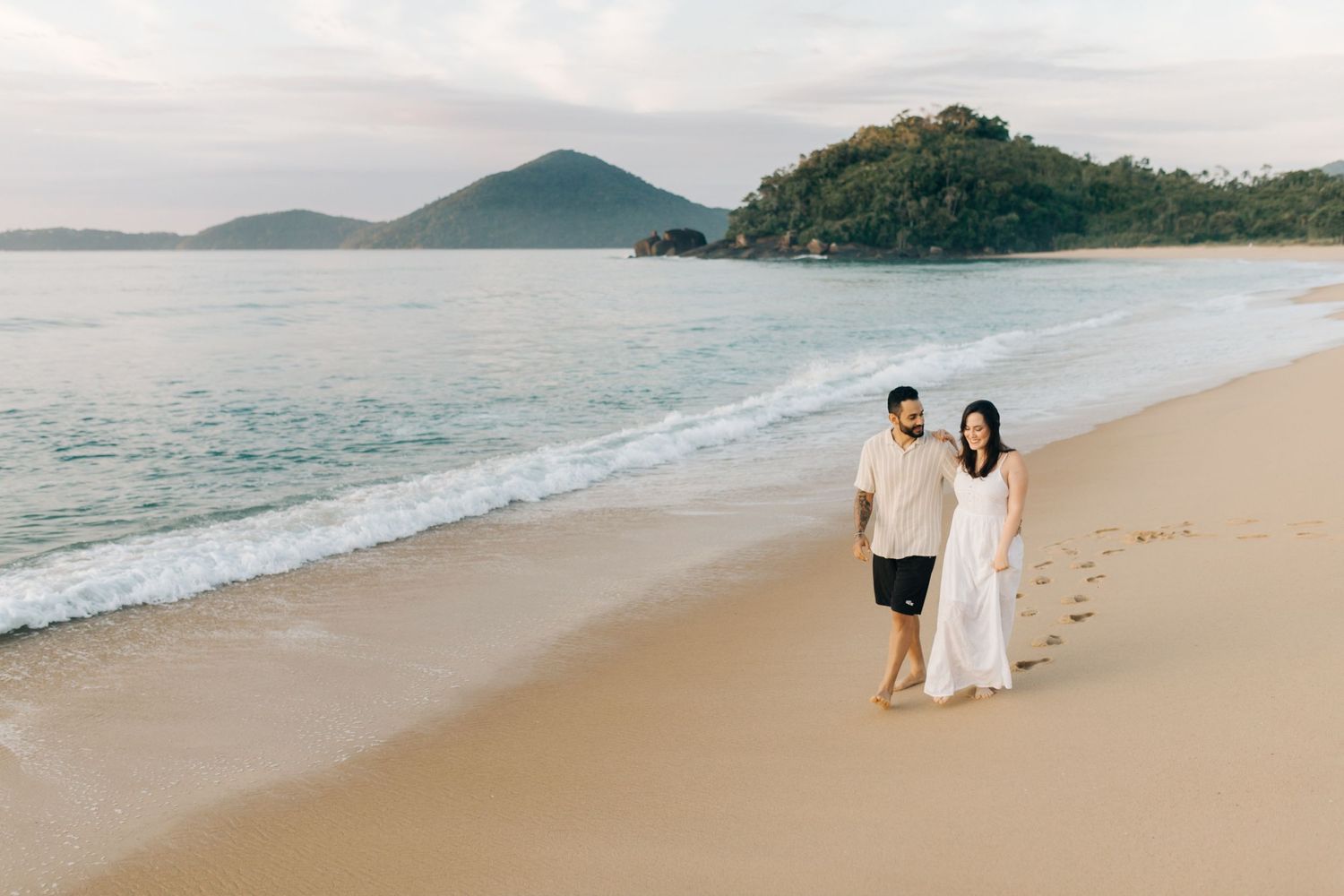 A couple walks along a pristine beach with mountains visible in the distance.