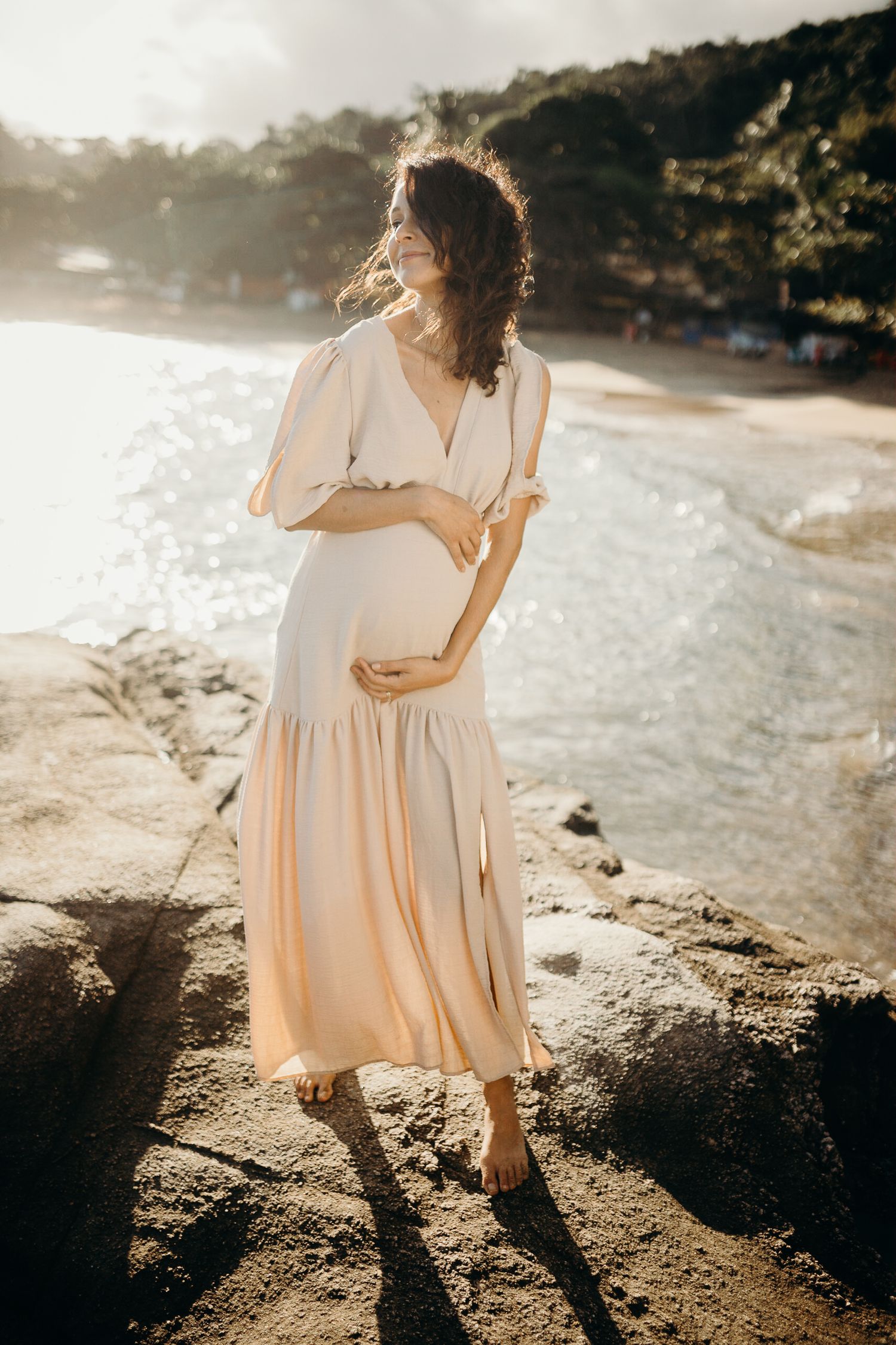 A person in a flowy beige dress poses on a rocky beach at sunset, creating long shadows on the ground.