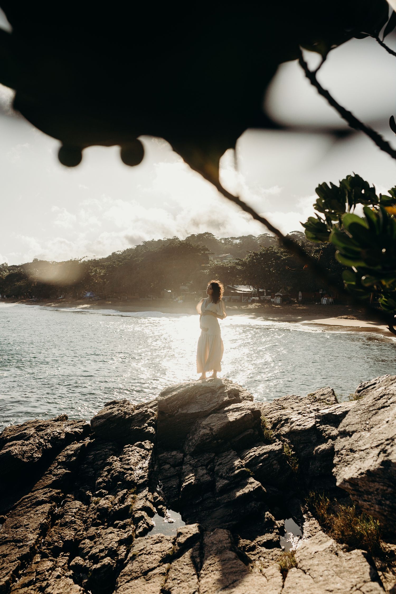Figure in white dress stands on coastal rocks overlooking tropical beach at sunset.