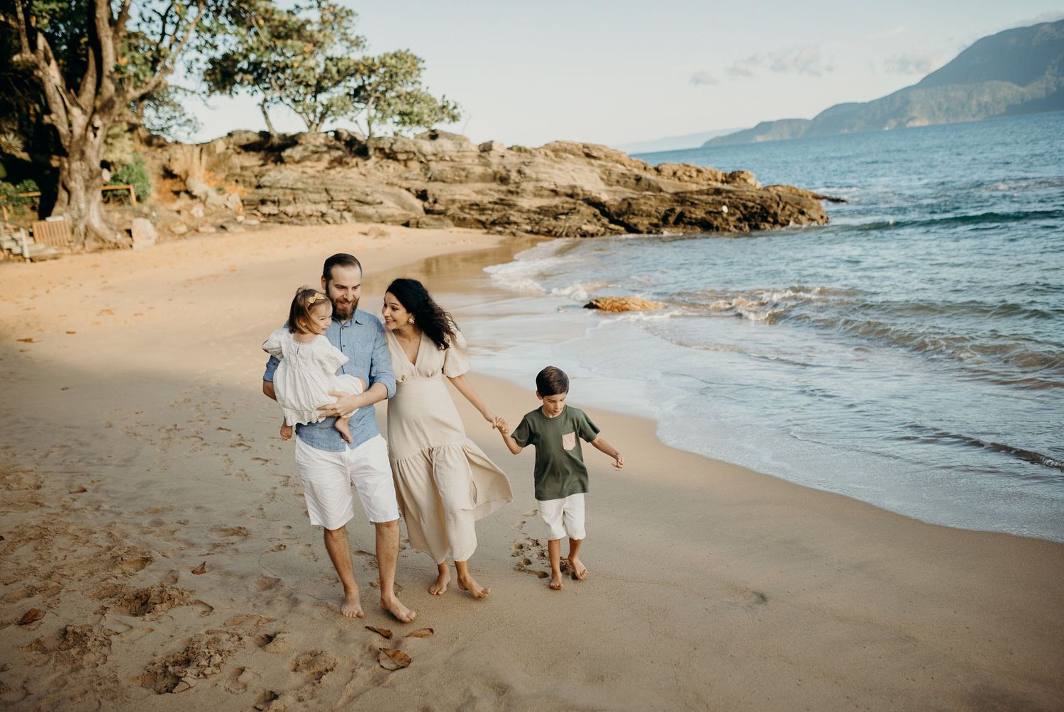 A family enjoys a beach walk together along the shoreline with mountains in the background during sunset.