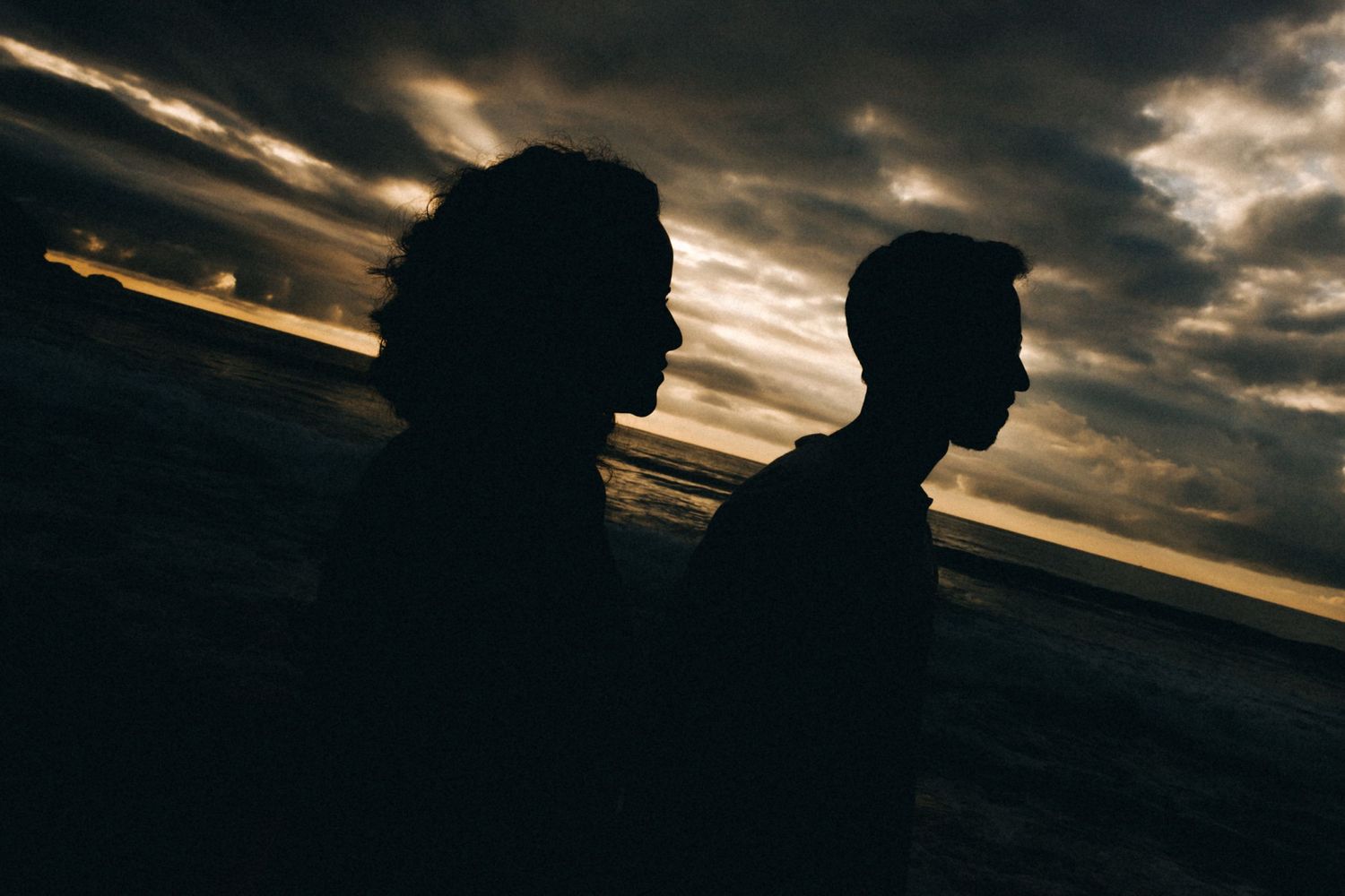 Silhouetted figures stand against a dramatic sunset sky at the beach.