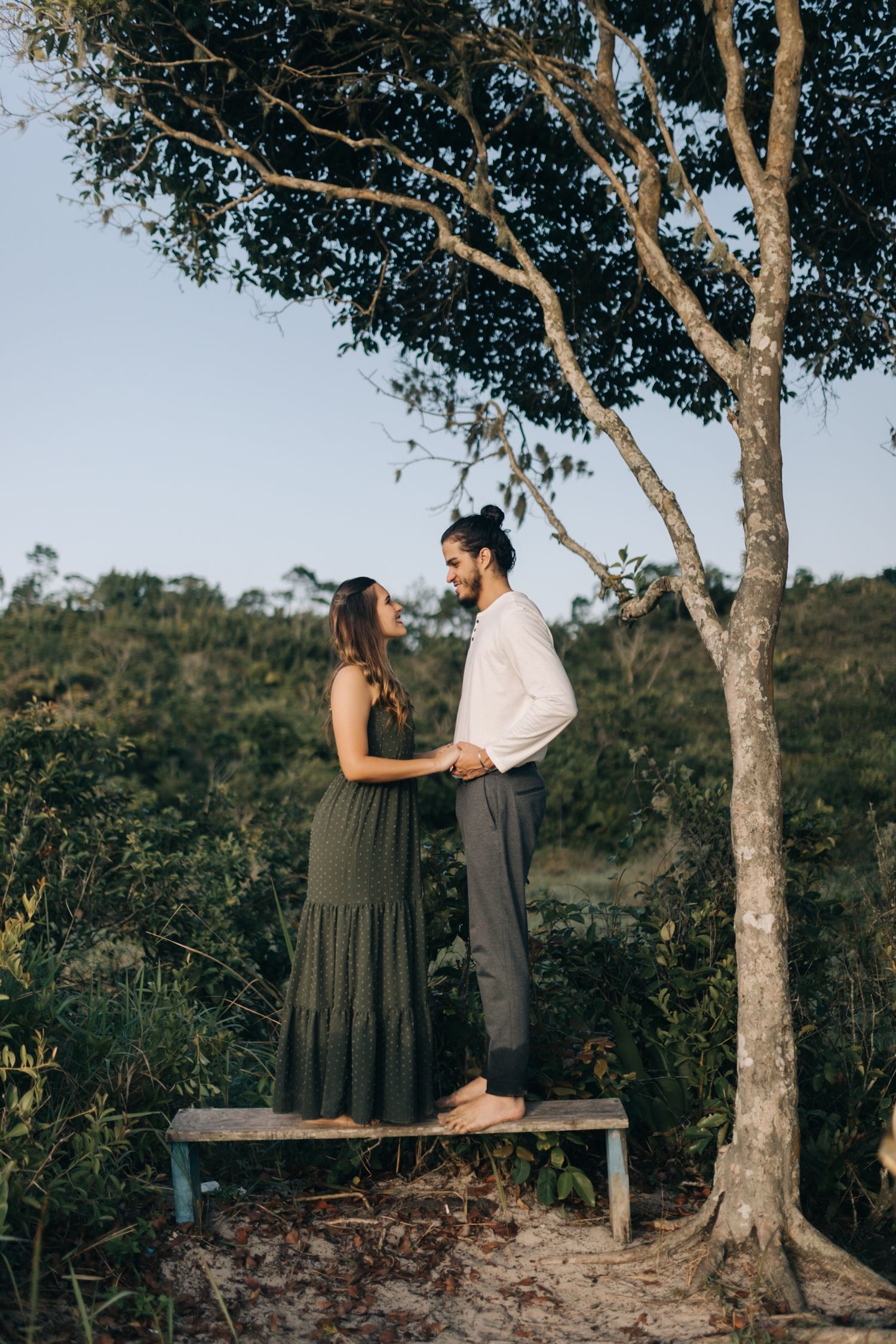 Couple posing on wooden bench surrounded by tall trees in natural setting.