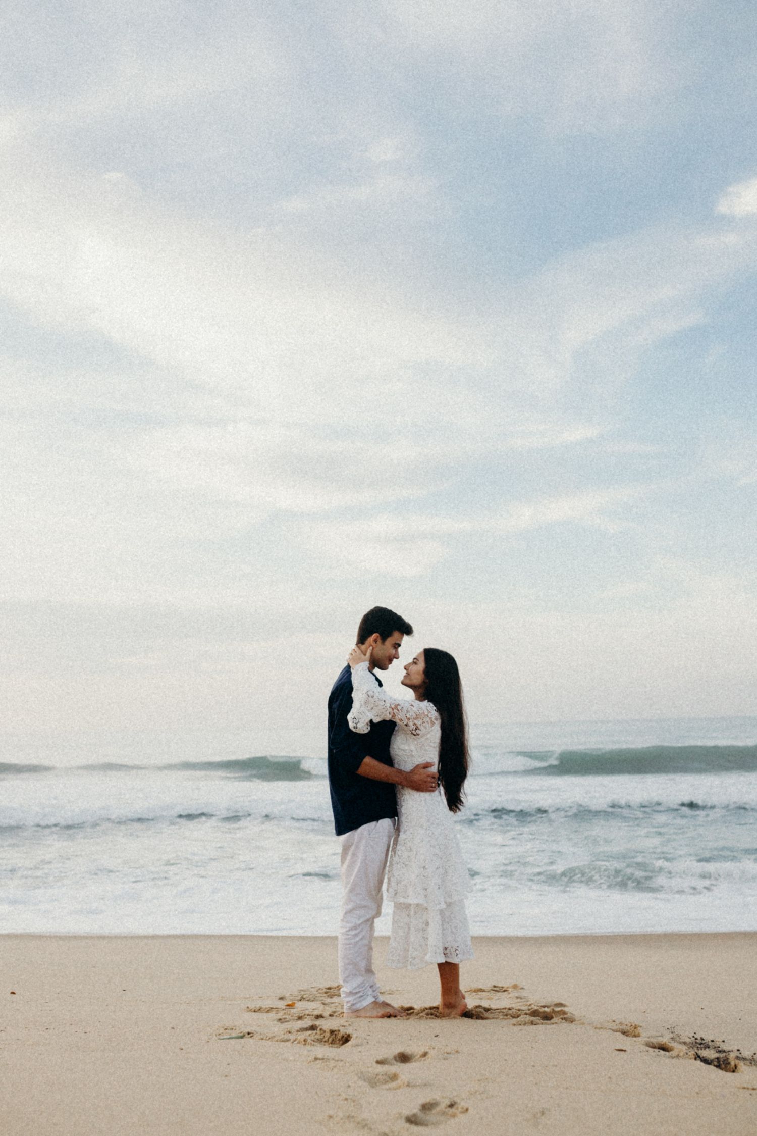 Romantic couple embracing on a beach with white lace dress and navy shirt against an ocean backdrop.