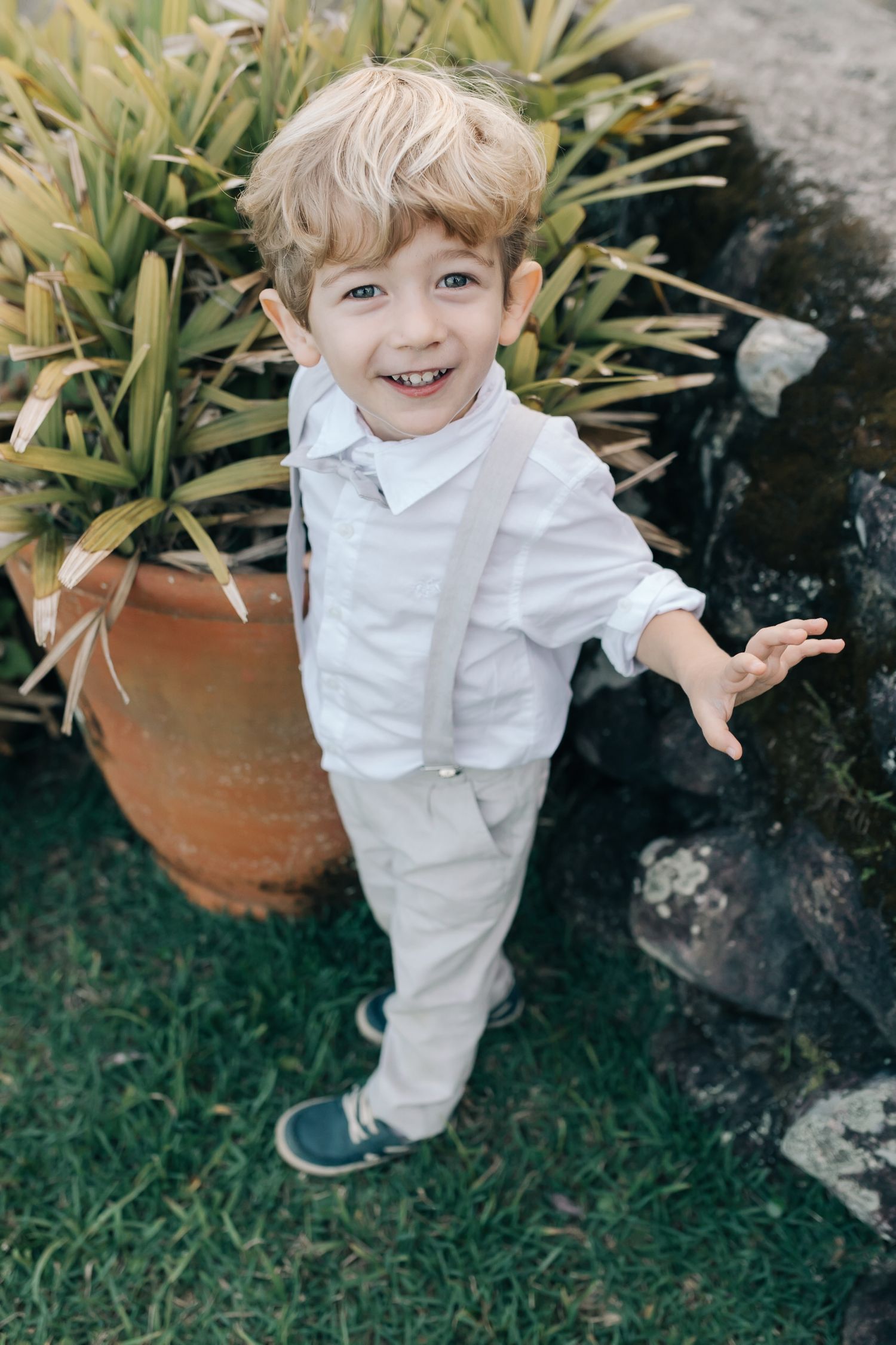 A toddler in an all-white outfit plays near potted plants in a garden setting.