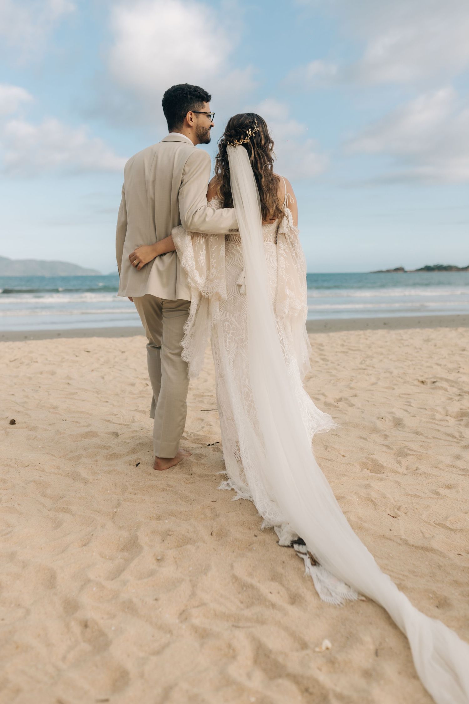 Back view of bride and groom embracing on sandy beach with long flowing veil.
