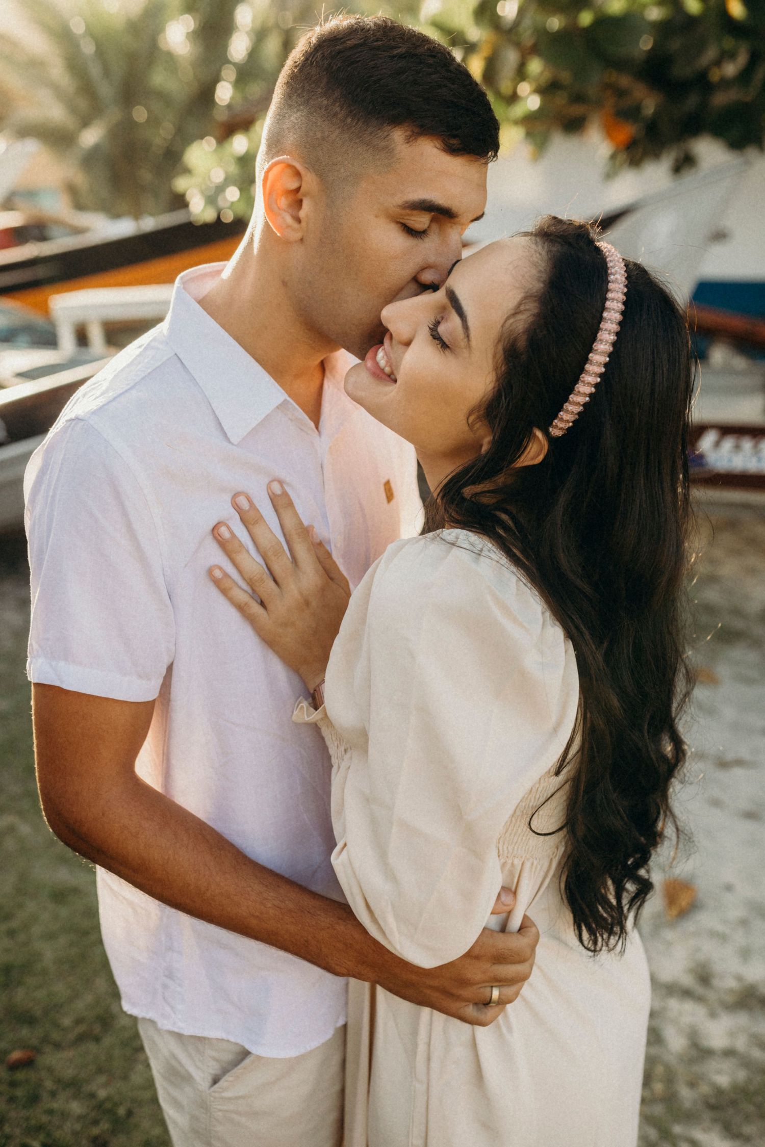 A tender moment between a couple in white outfits standing among boats and palm trees.