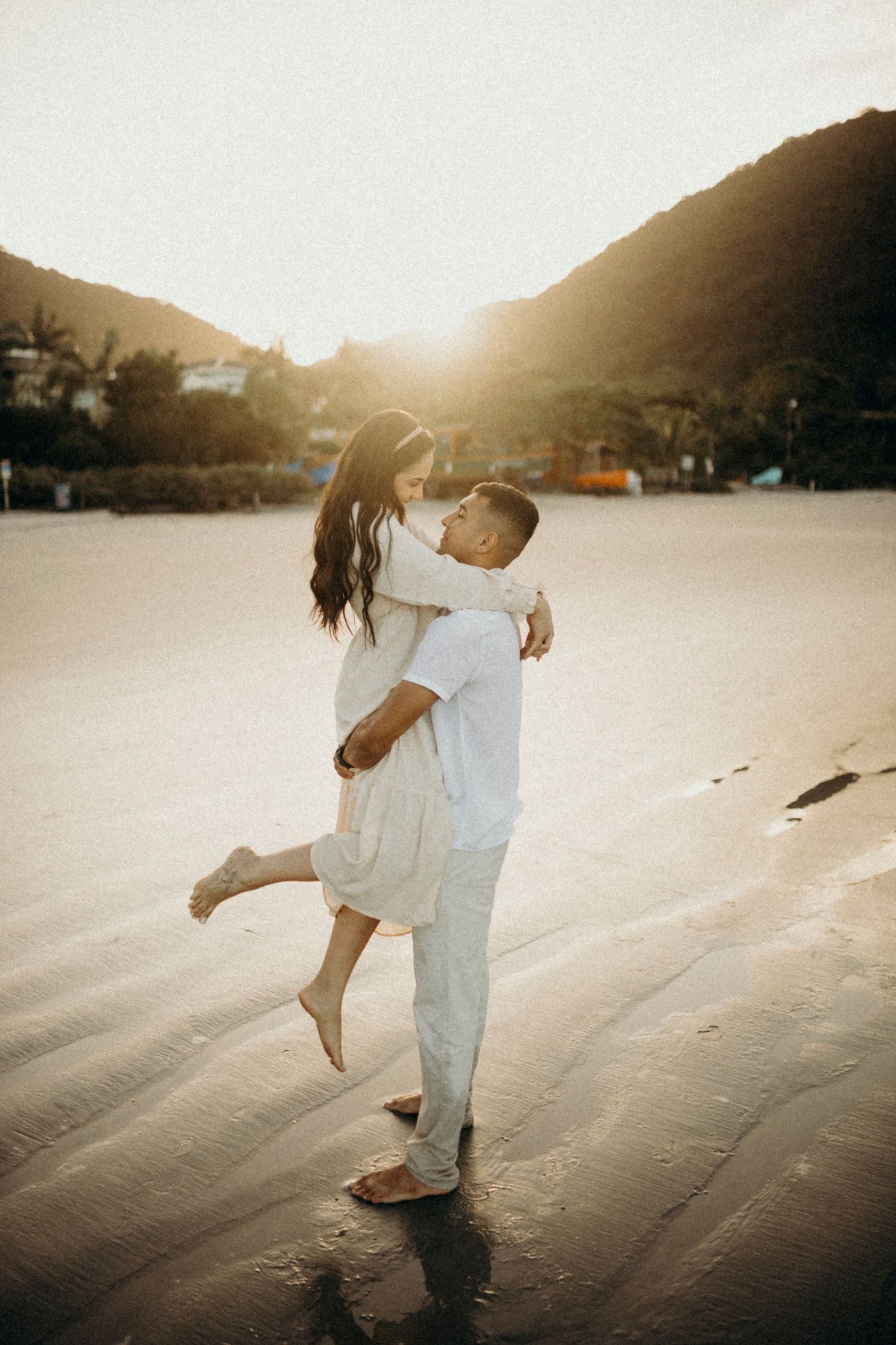 Couple in light clothing embraces on sandy beach at golden hour with mountains in background.