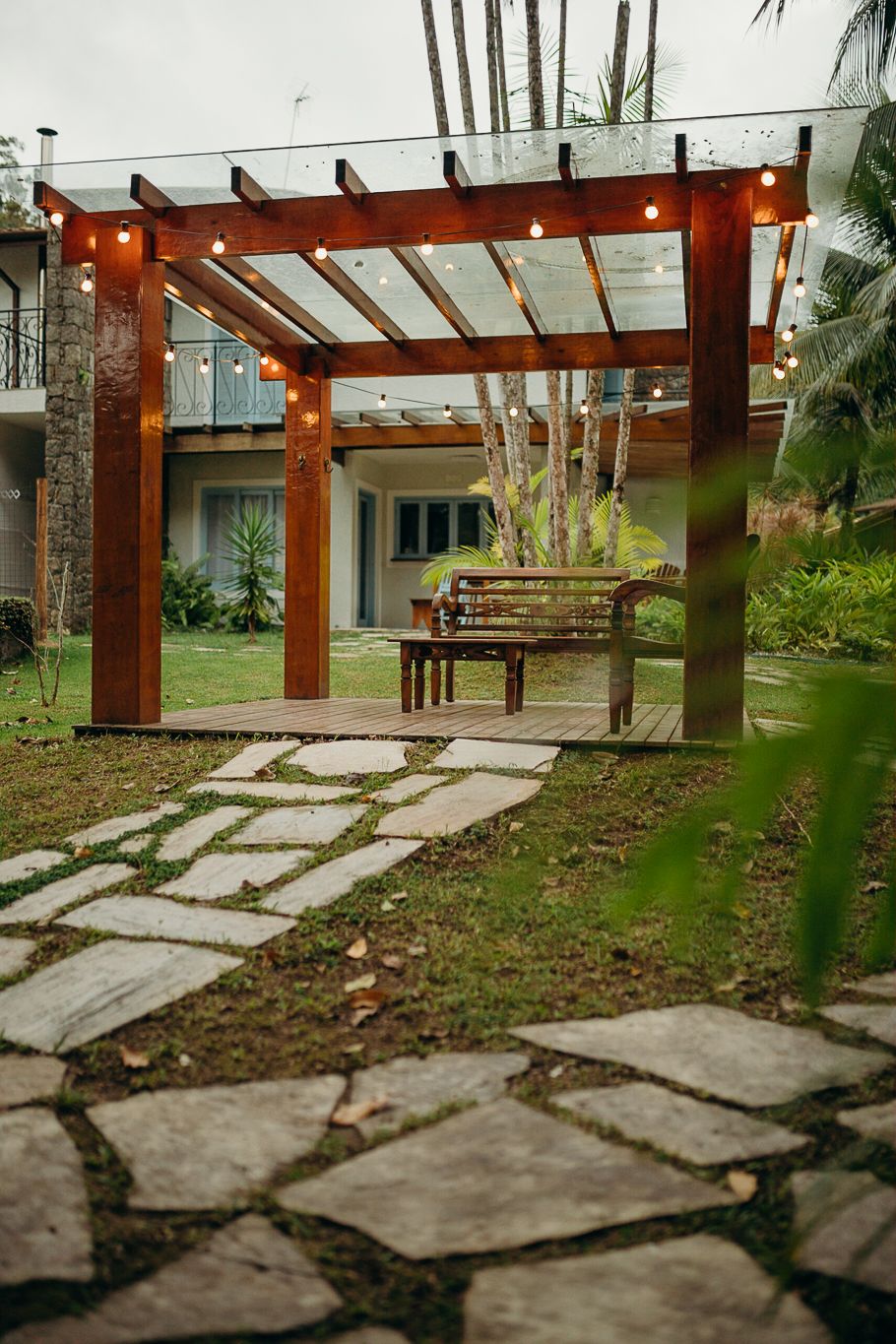 Stone pathway leading to wooden pergola with string lights in lush tropical setting.