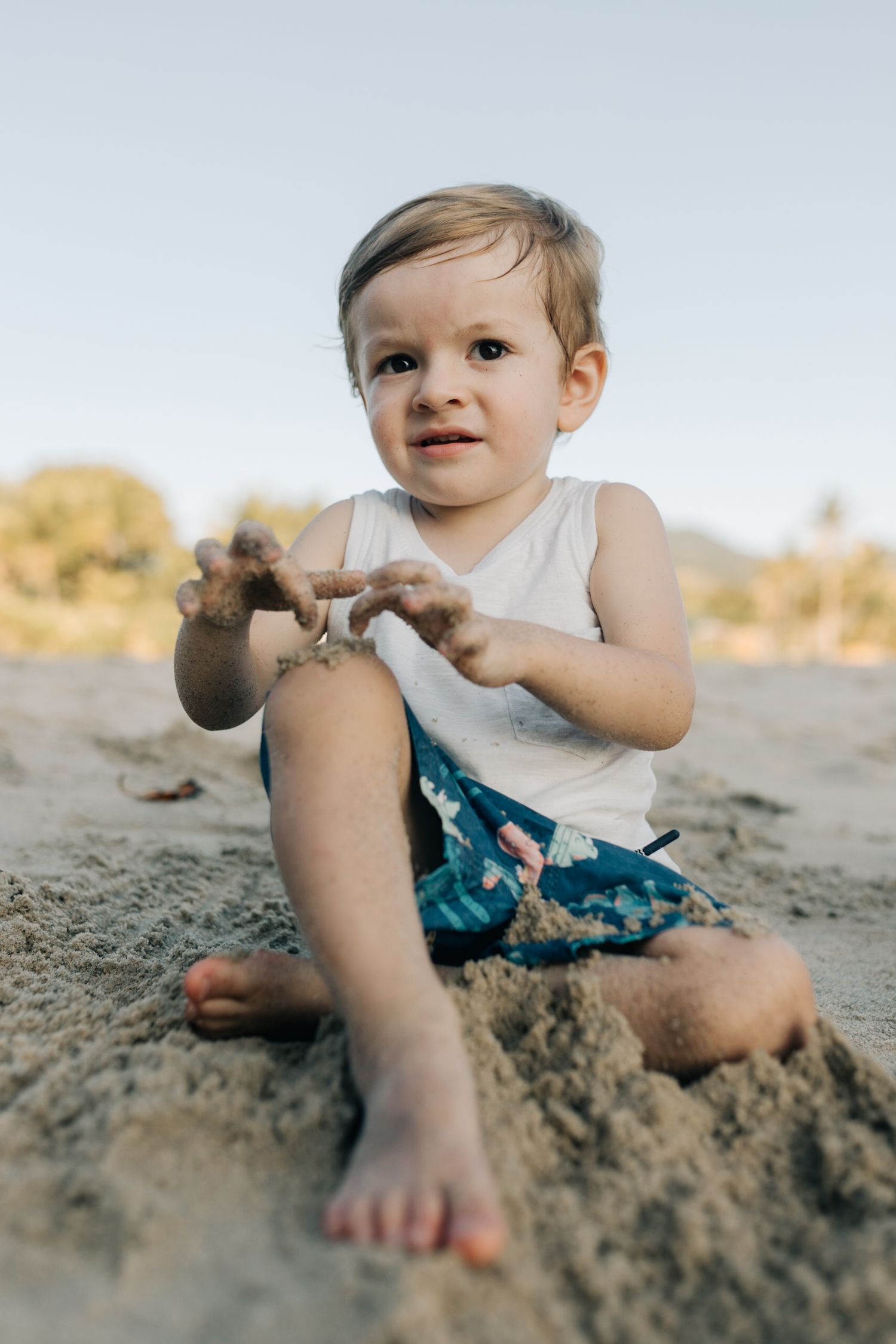 A toddler in a white tank top and blue shorts plays in the sand at the beach.
