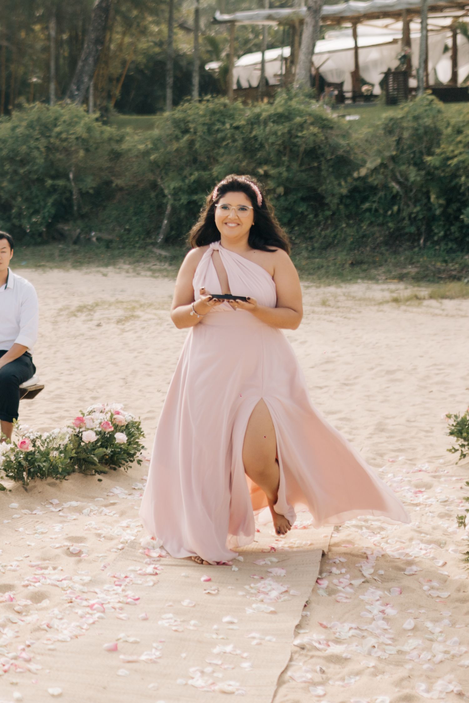 A bridesmaid in a flowing pink dress walks down a sandy aisle decorated with rose petals.