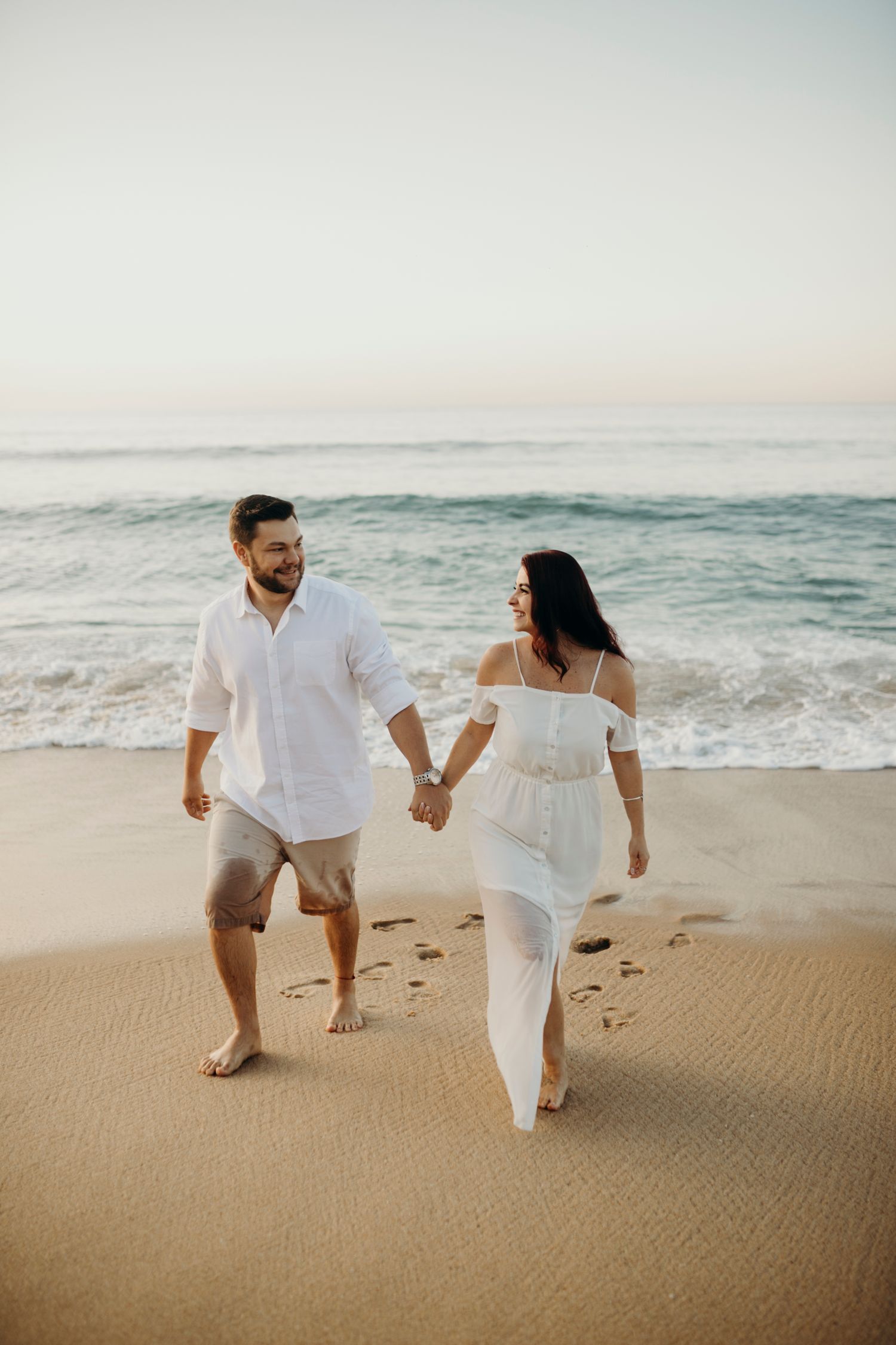A couple holding hands while walking barefoot along a scenic beach shore.