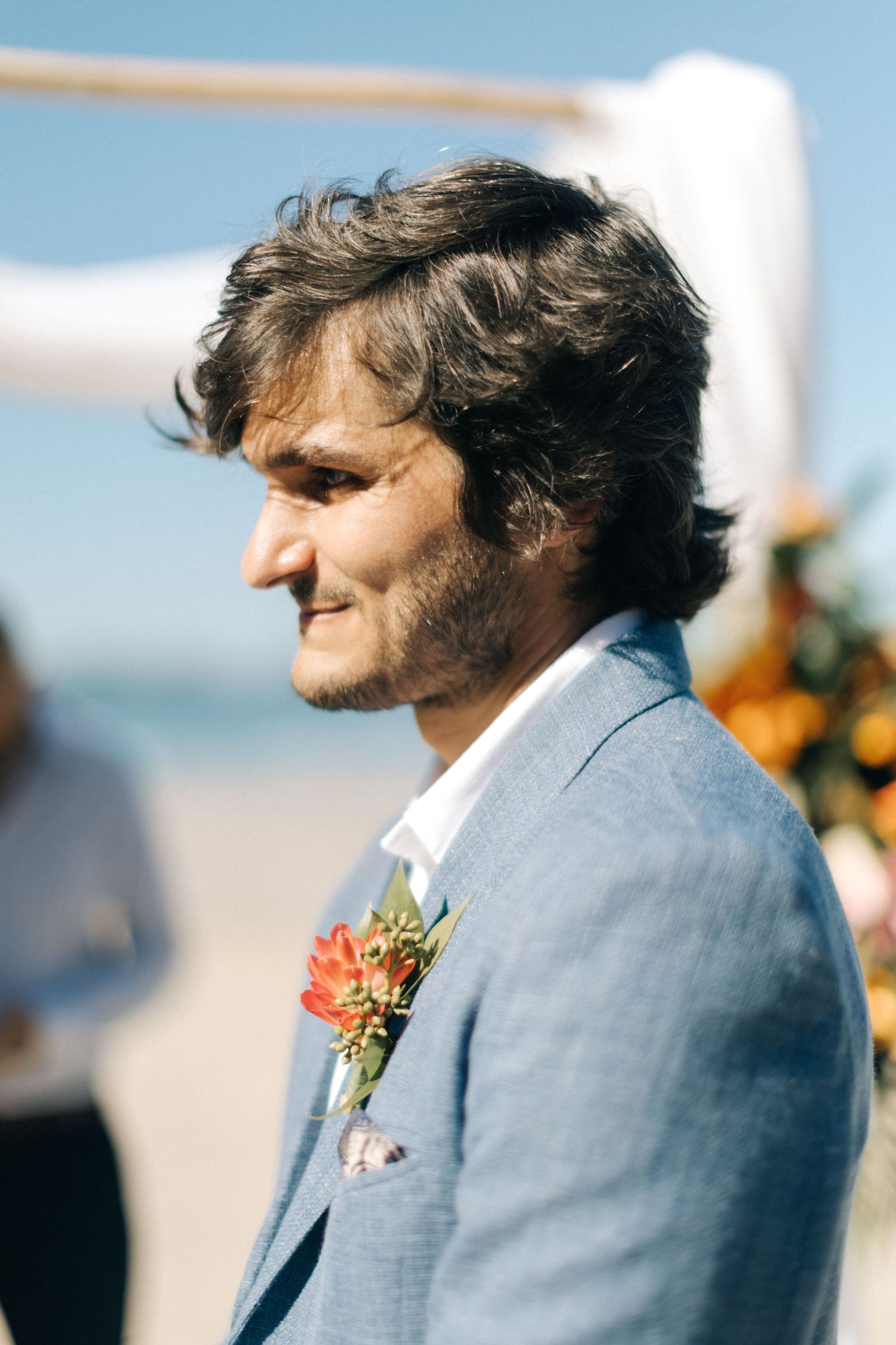 A groom in a light blue suit standing on a beach looks to the side during a wedding ceremony.