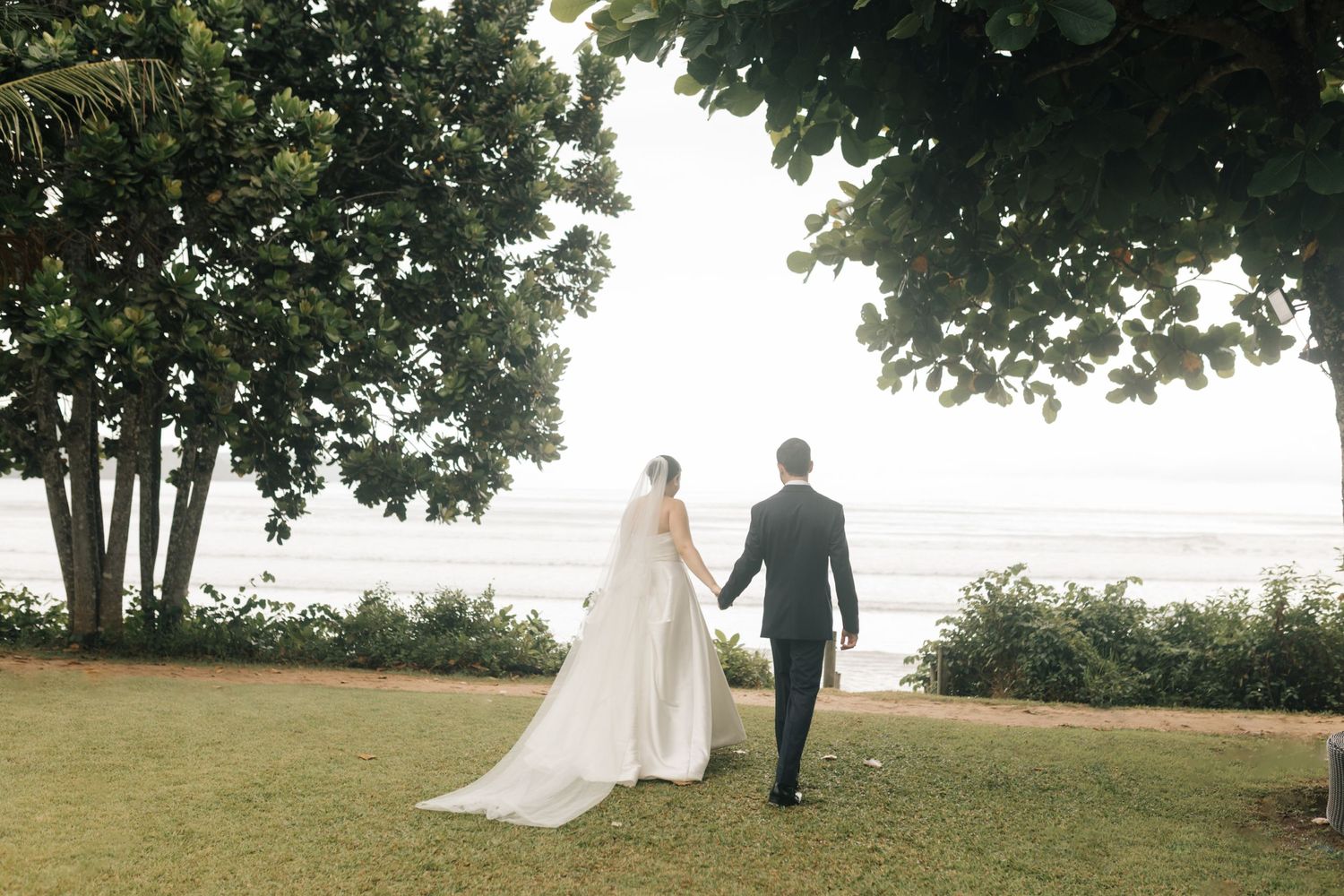 A bride and groom walk hand in hand on a grassy lawn with trees framing the ocean view.