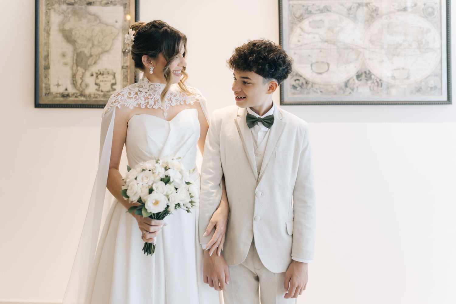 Elegant wedding couple in white attire posing against neutral wall with framed artwork.