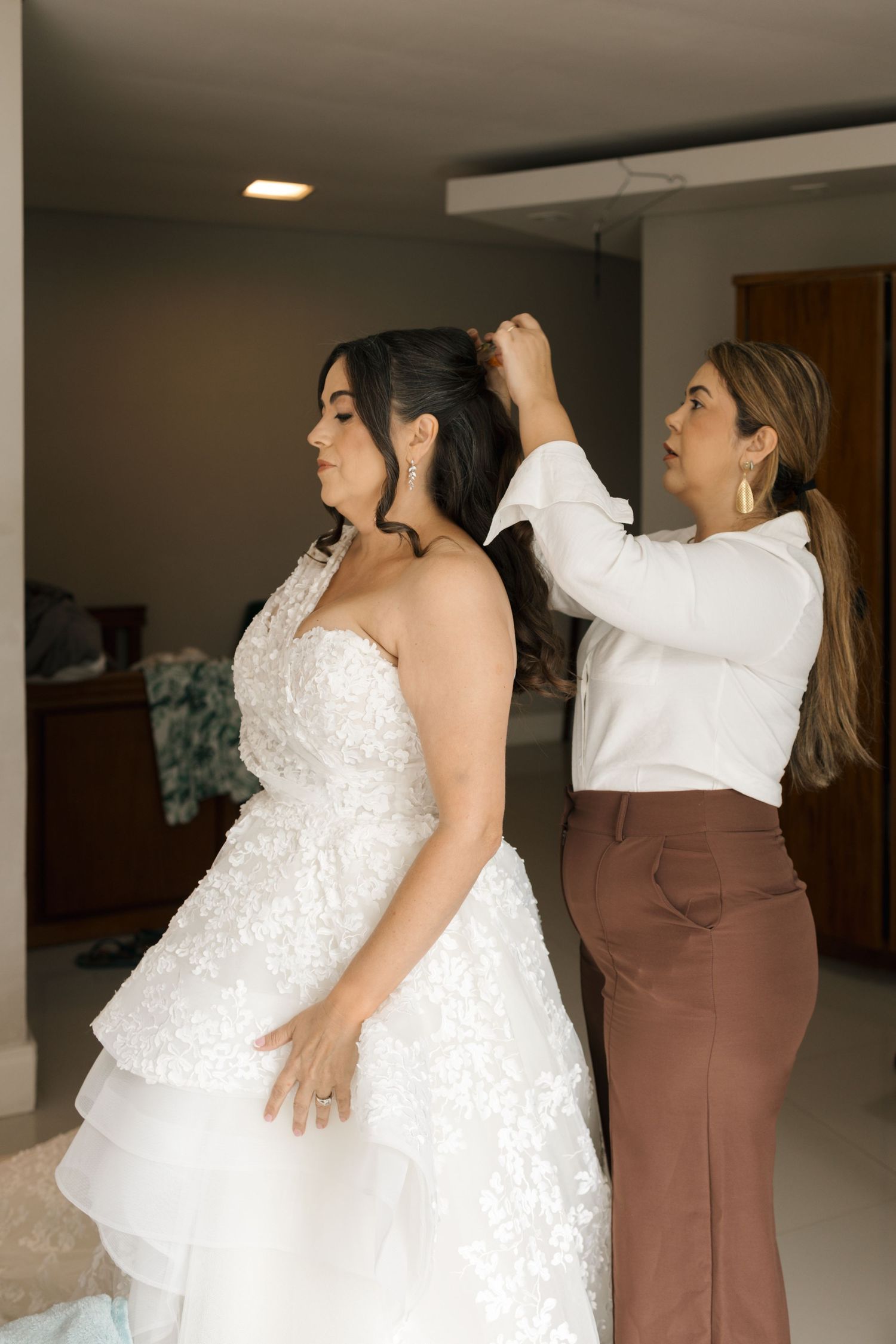 Bride in a white lace wedding dress being helped by attendants in brown dresses during wedding preparations.