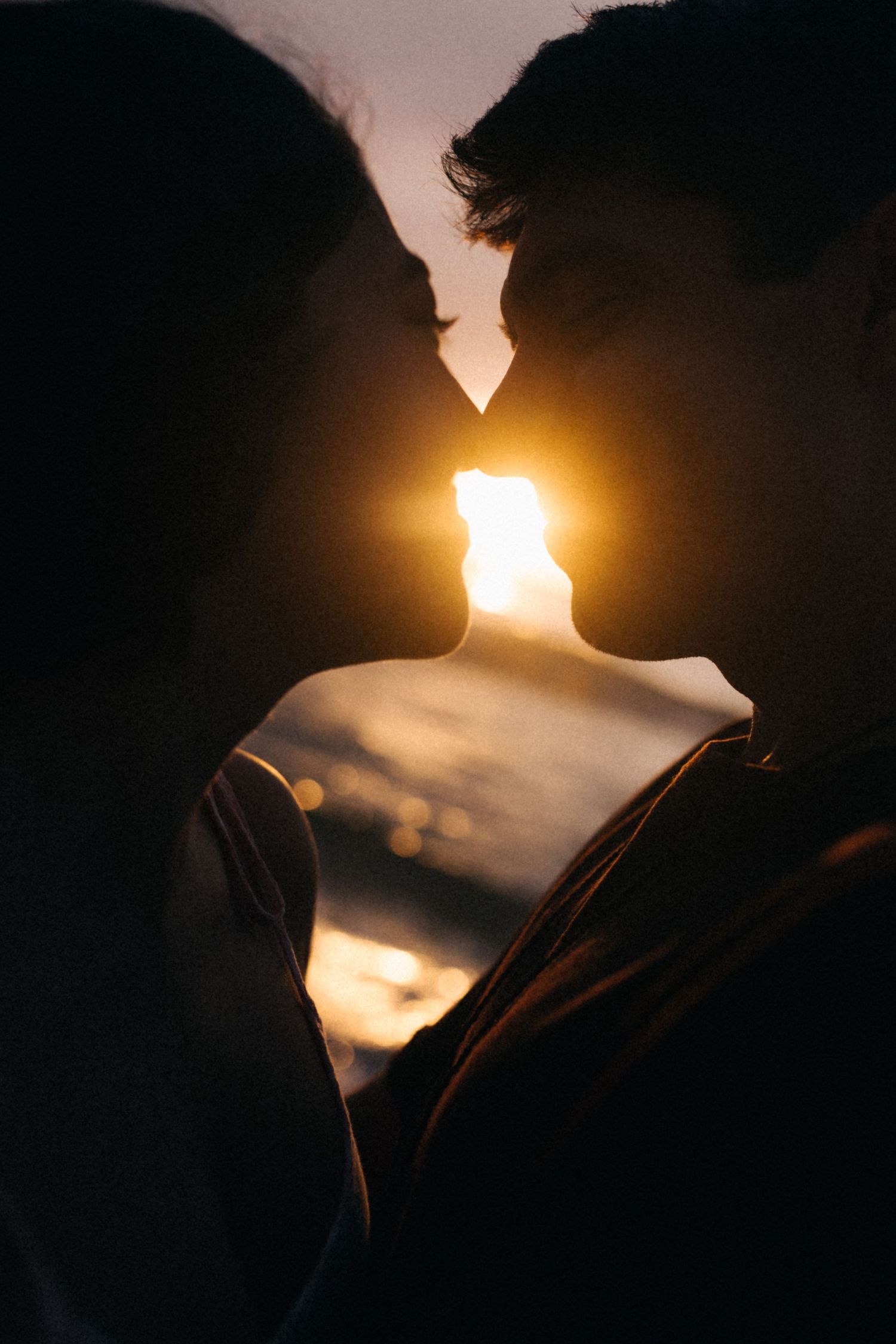 Casal em silhueta em pose íntima contra o pôr do sol dourado criando um efeito romântico retroiluminado na água.