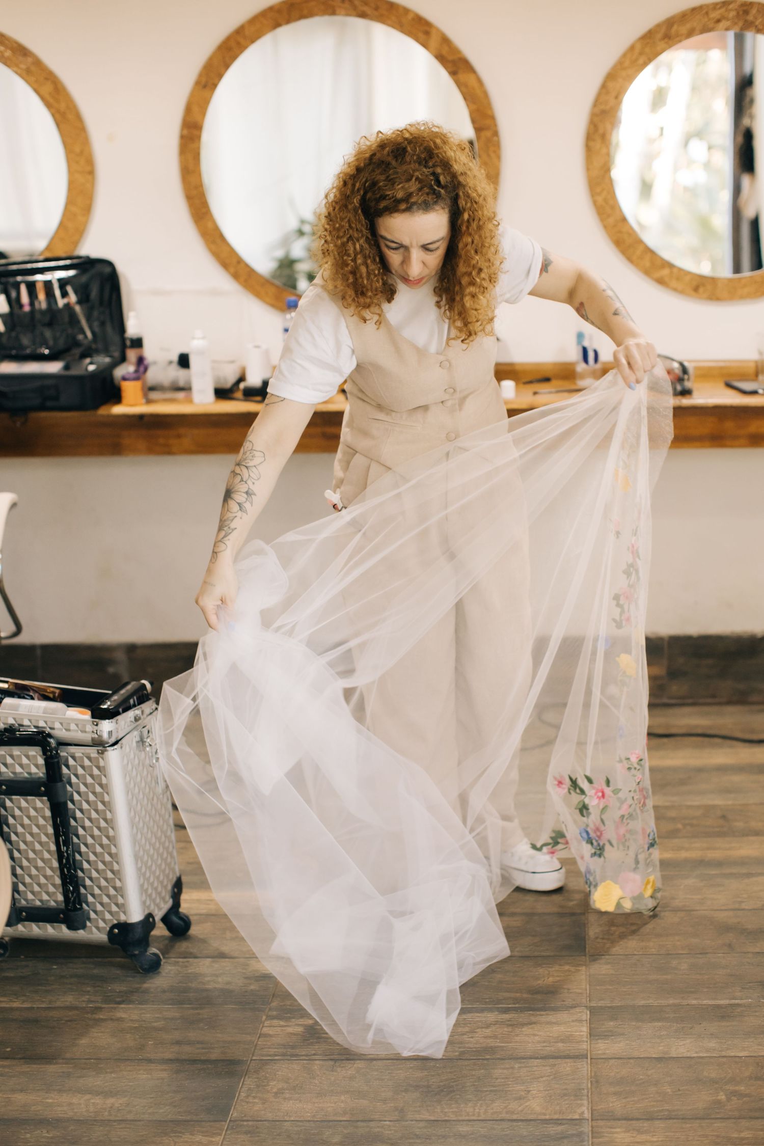 Stylist in beige outfit arranging white tulle wedding veil in salon with round mirrors.