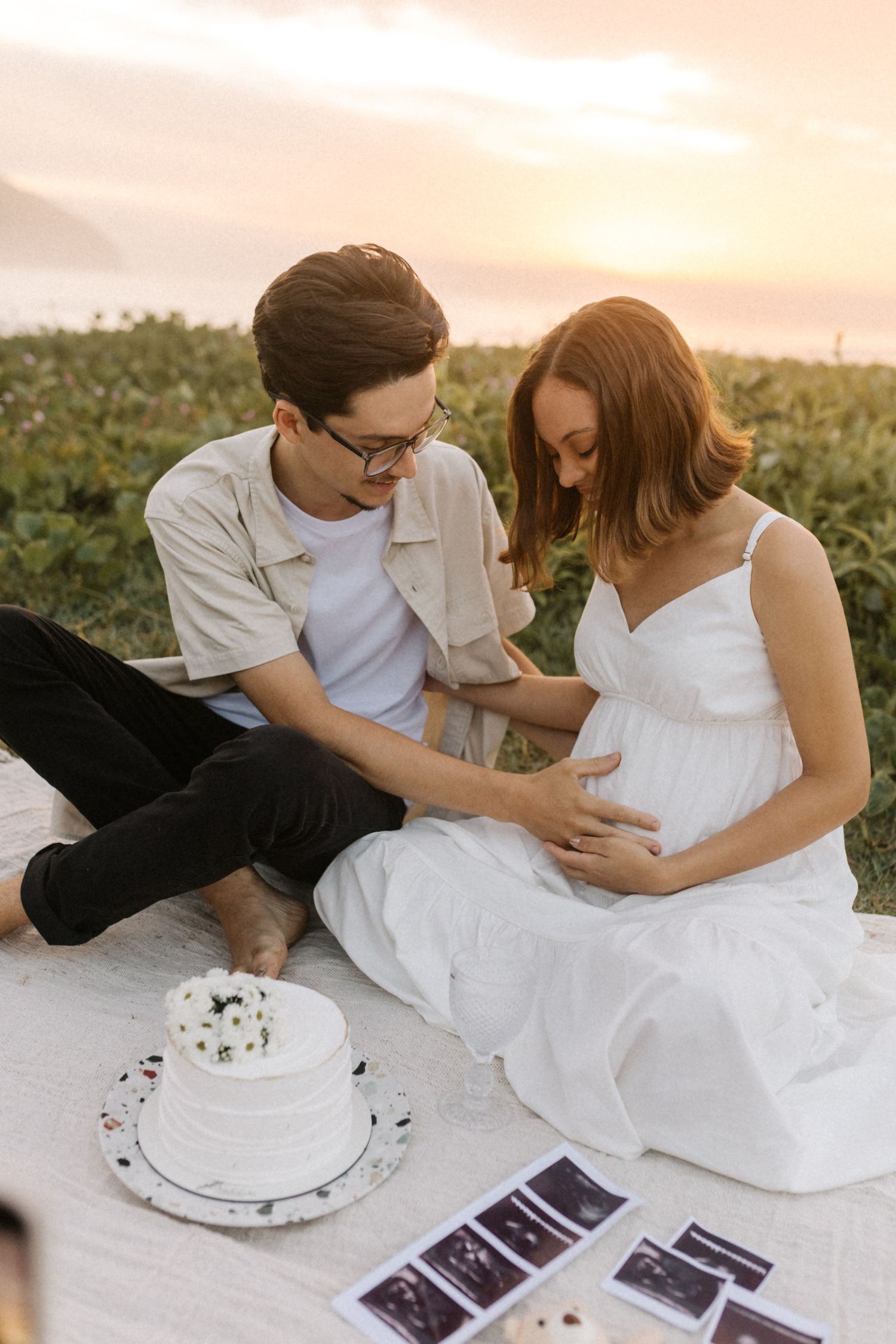 An expectant couple looks at ultrasound photos during a sunset picnic with a white cake.