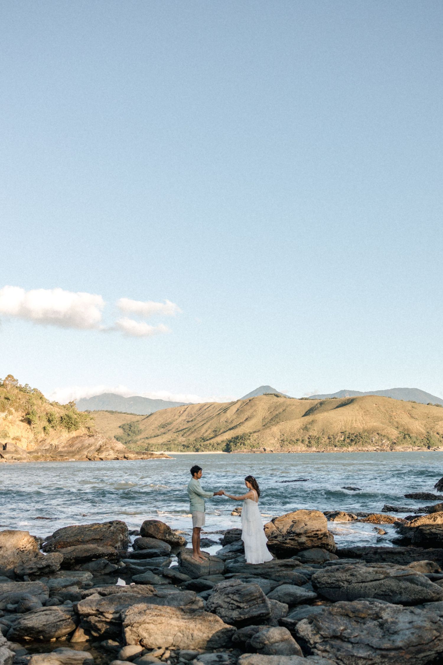 Wedding couple stands on rocky coastline with mountains and ocean in the background.