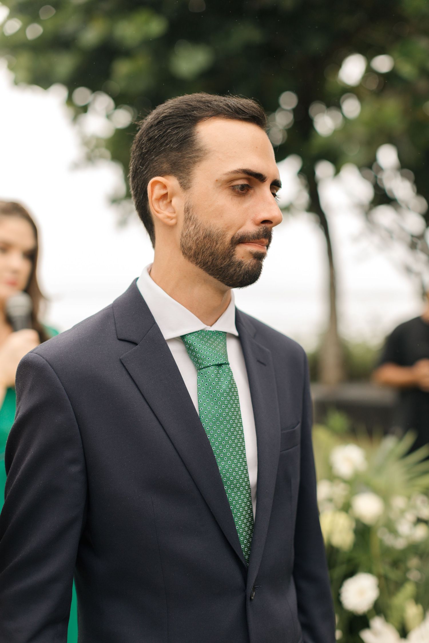 A well-dressed man in a navy suit with emerald green tie stands during an outdoor ceremony.