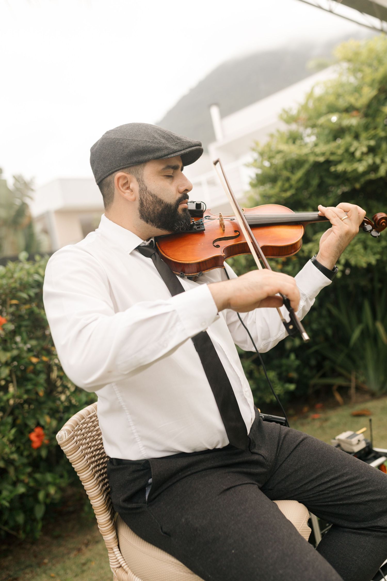 Musician playing violin outdoors while seated in casual attire with cap.