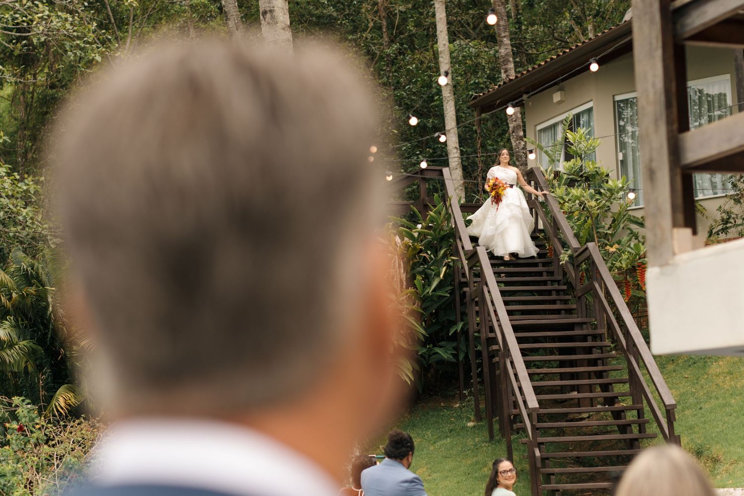Outdoor wedding venue with string lights and wooden stairs visible through trees.