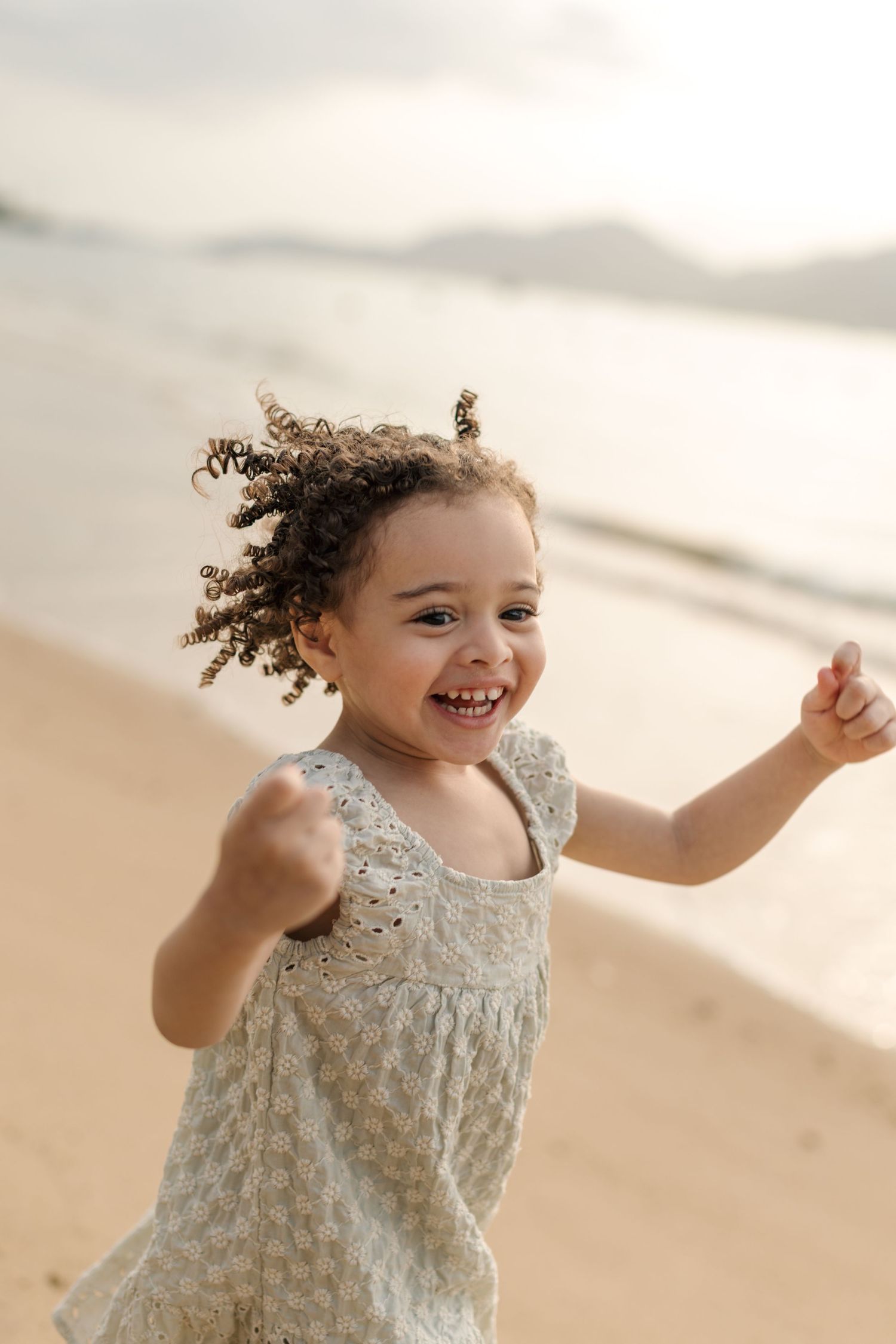 A joyful child with curly hair plays and dances on a sandy beach at sunset.