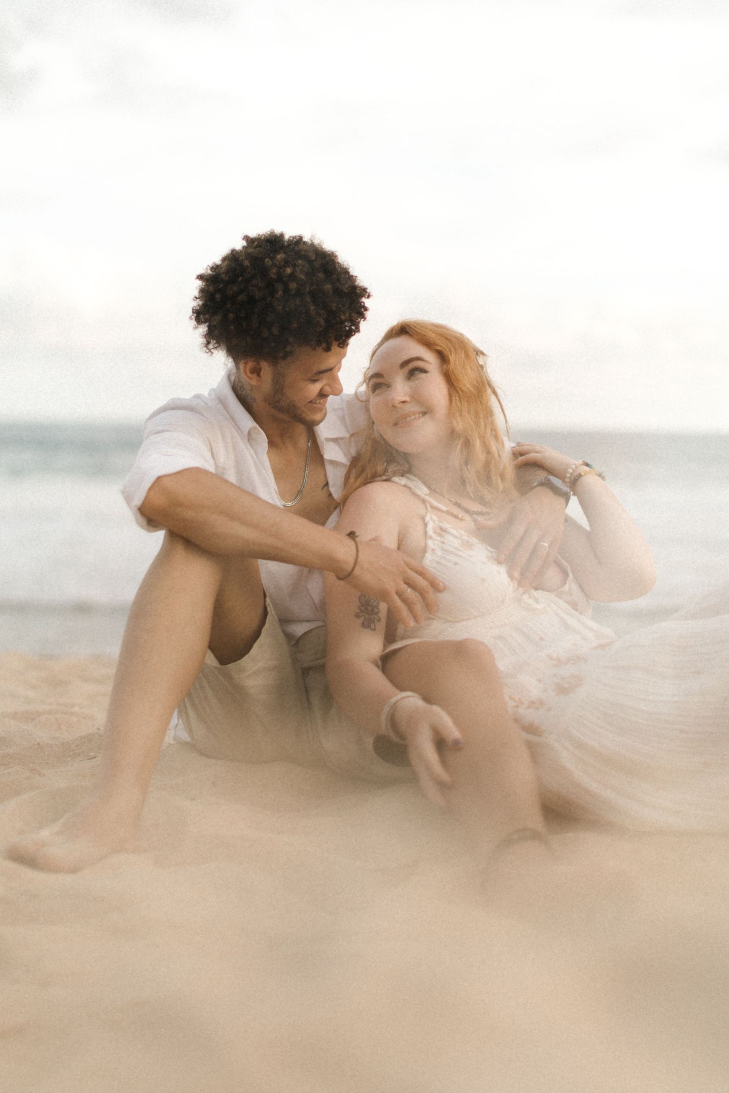 A romantic couple embraces on a sandy beach with ethereal lighting and flowy fabric creating a dreamy atmosphere.