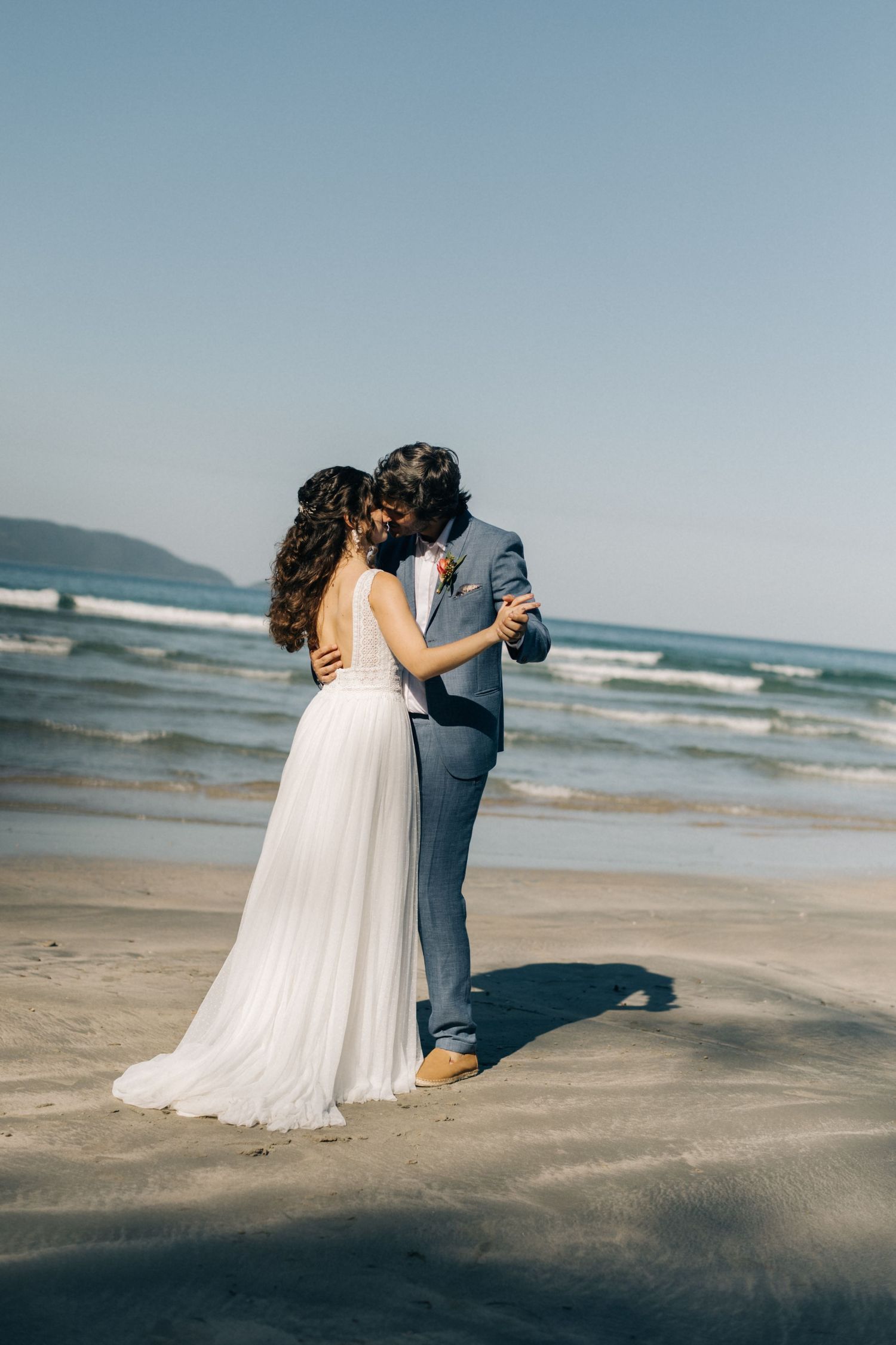 A couple in wedding attire dances together on a beautiful sandy beach with ocean waves in the background.