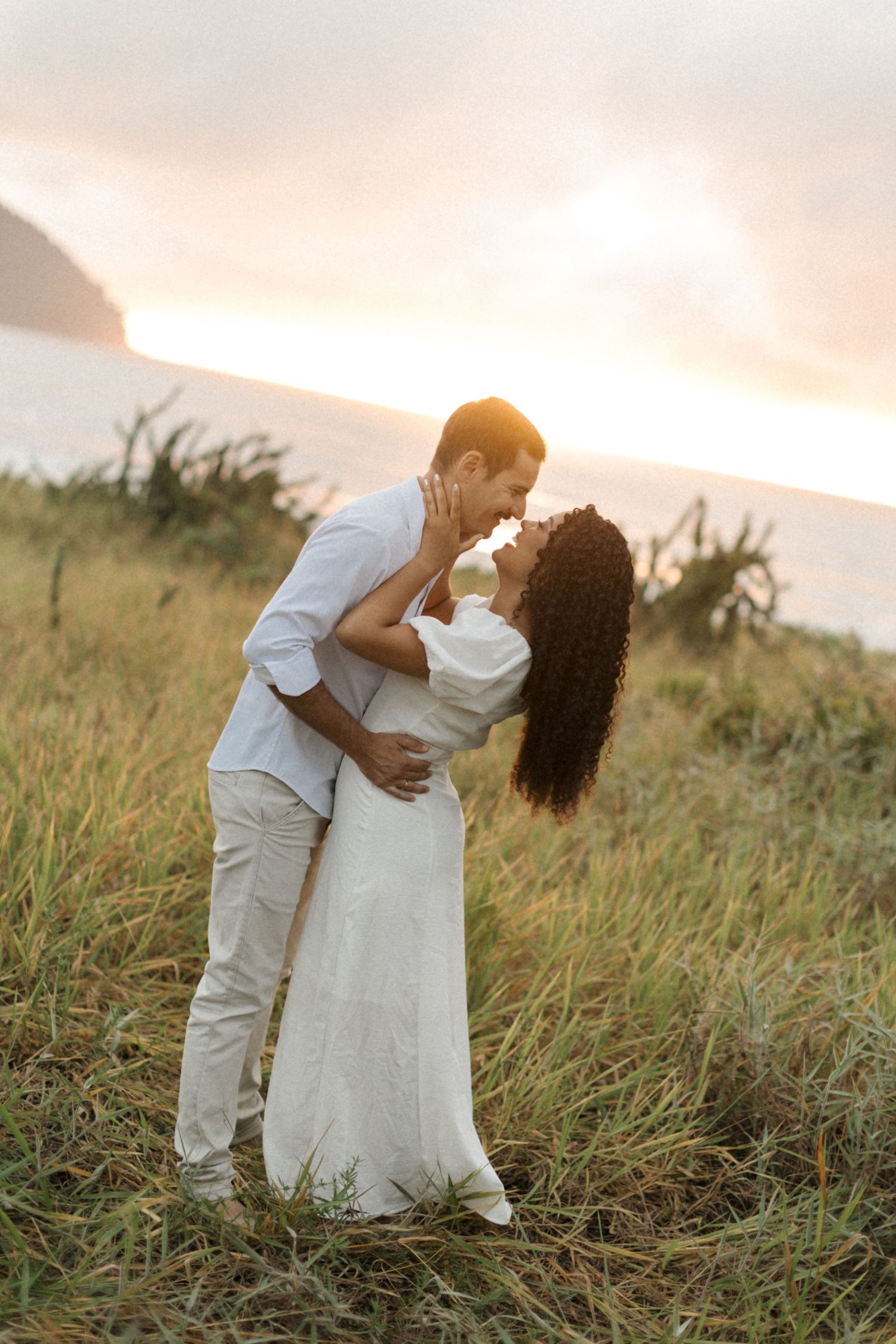 A romantic couple embraces in a grassy field during golden hour sunset with mountains in the background.