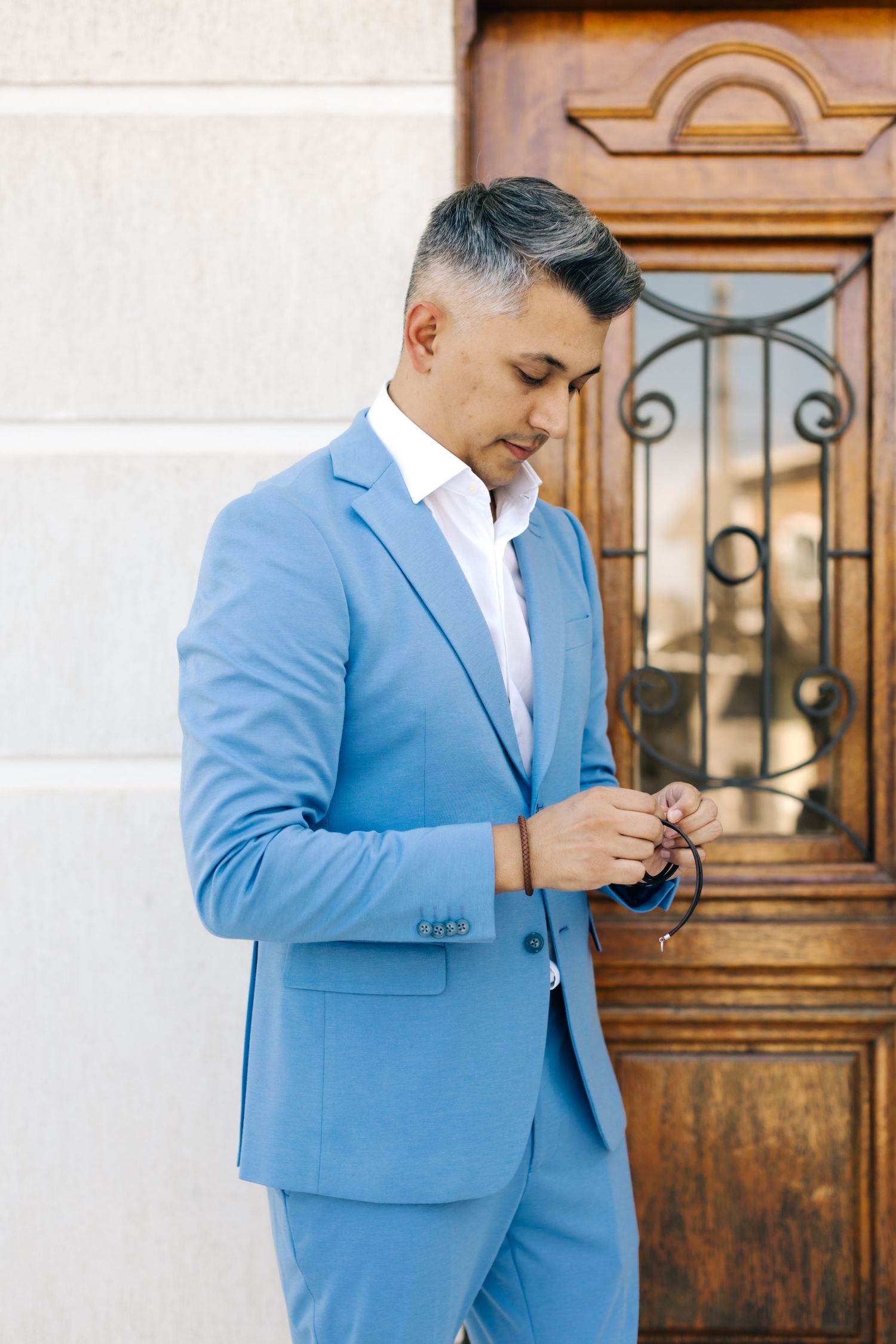 Man in light blue suit checking phone outside elegant wooden doorway.