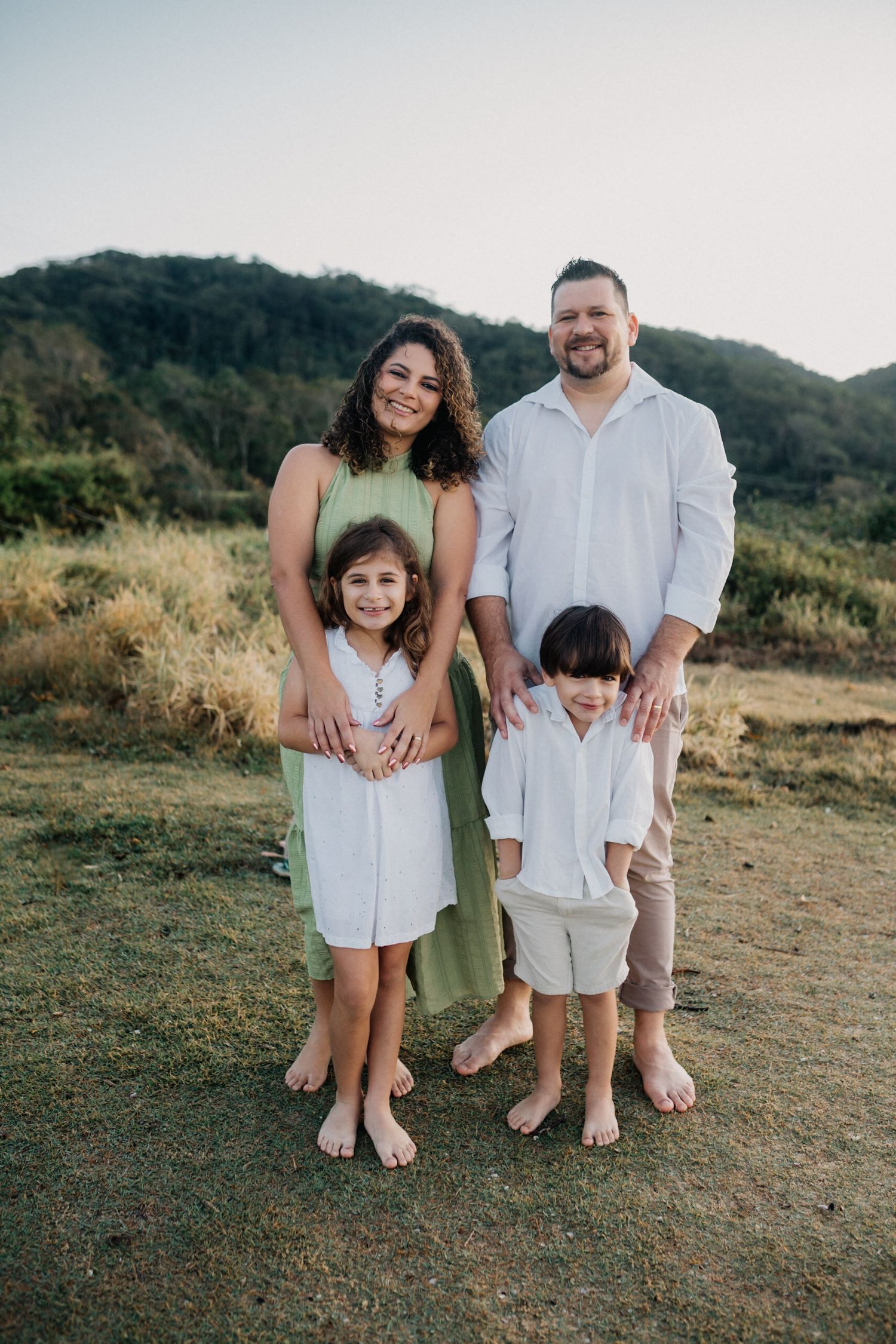 Family sitting together barefoot on grassy hillside wearing coordinated white and beige outfits.
