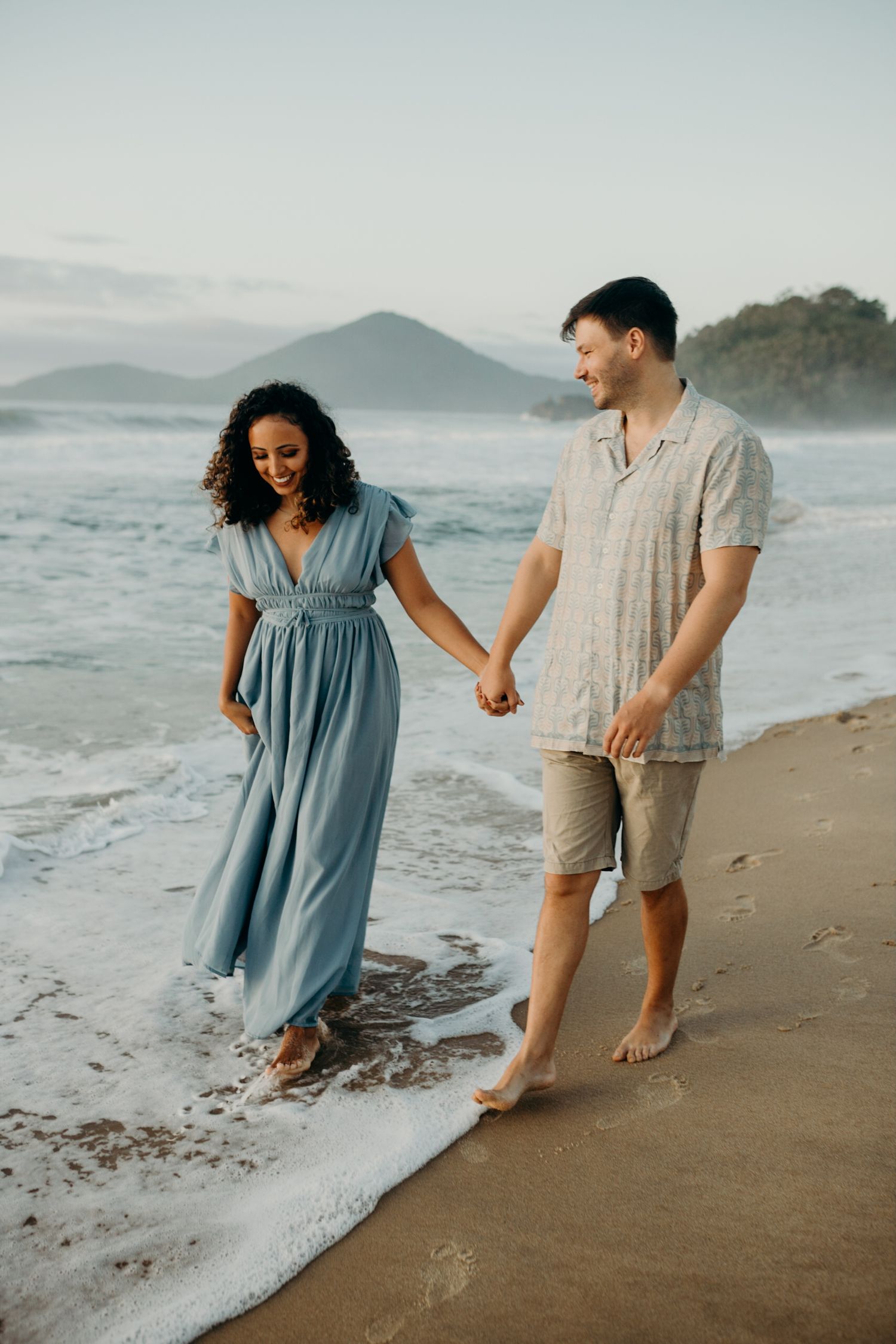 Playful barefoot couple walking at water's edge on sandy beach with mountains in background.