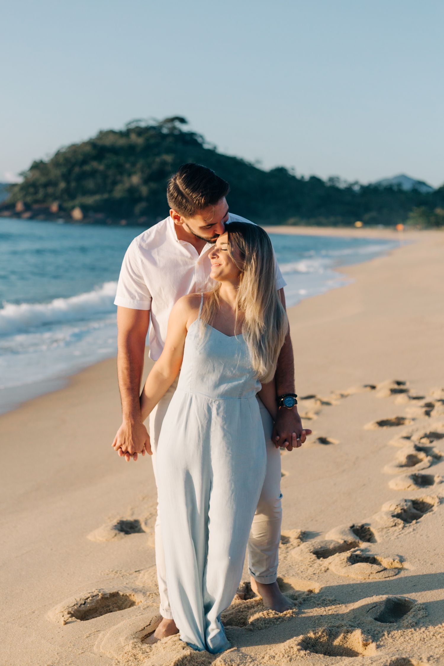 Romantic embrace between couple in white outfits on sunny beach with mountains in background.